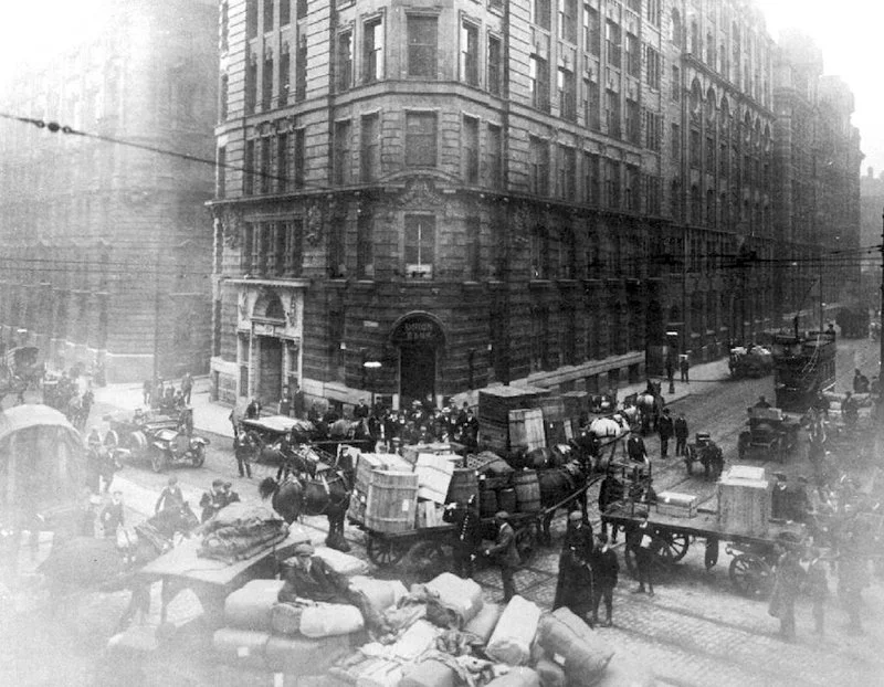 Early 20th-century city street scene with horse-drawn wagons, pedestrians, and a large multi-story building in the background.