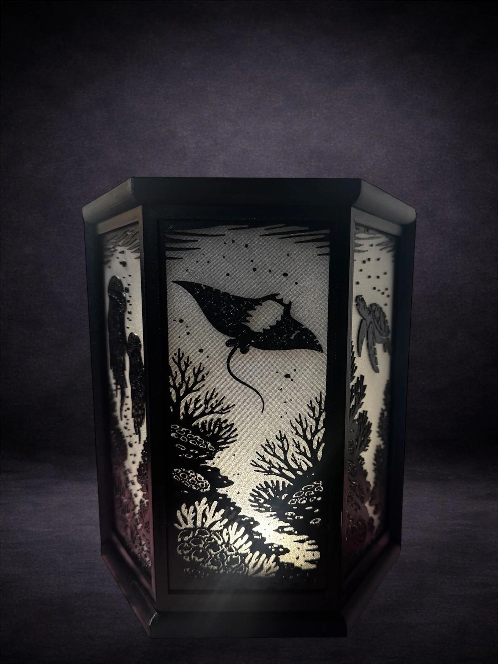 A decorative lantern with a black metal frame and intricately cut out panels depicting underwater scenes of coral, fish, and a flying manta ray against a dark background.