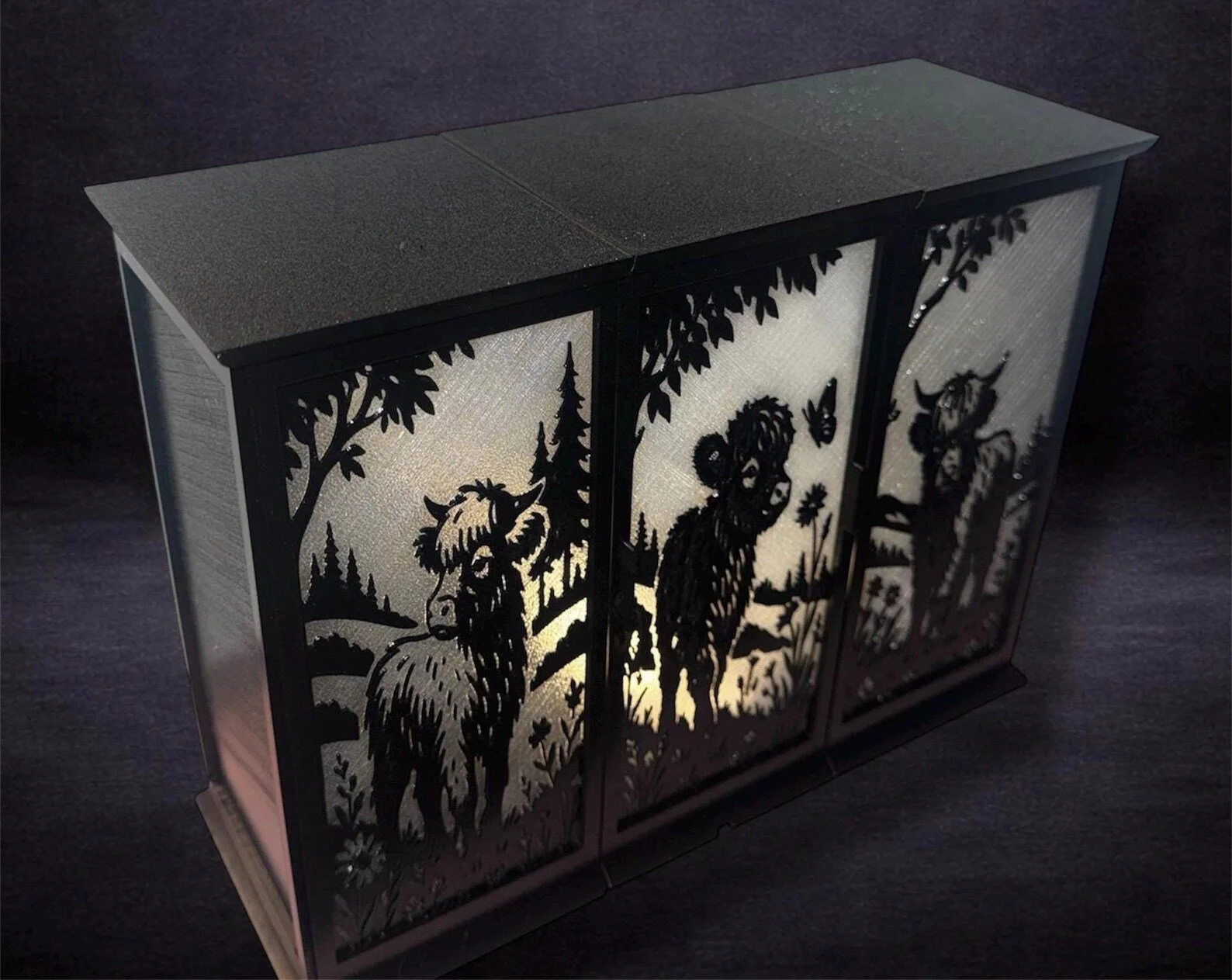 Decorative LED lantern with black silhouette cutouts of a moose and a bear in a forest scene, illuminated from within, against a dark background.