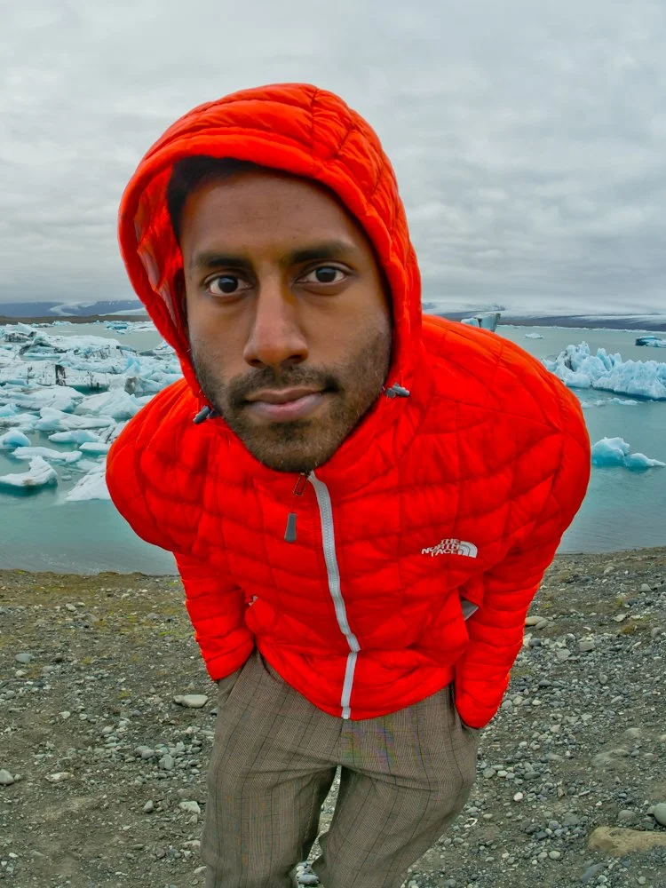 A man in a bright red jacket with a hood, standing outdoors near icebergs in a glacial landscape with a cloudy sky.