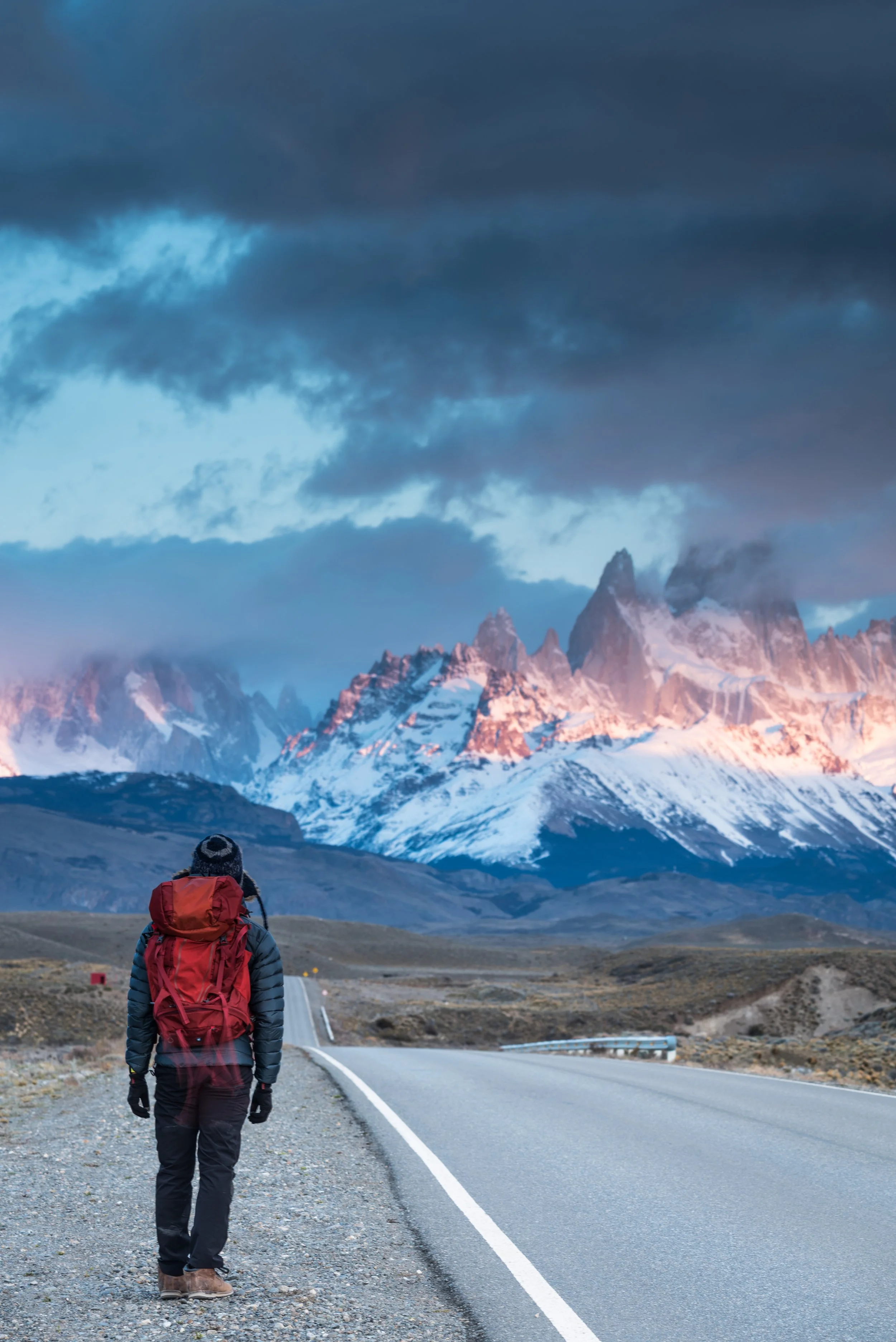 A hiker with a red backpack and winter jacket walking along a mountain road with snow-capped peaks in the background under cloudy skies.