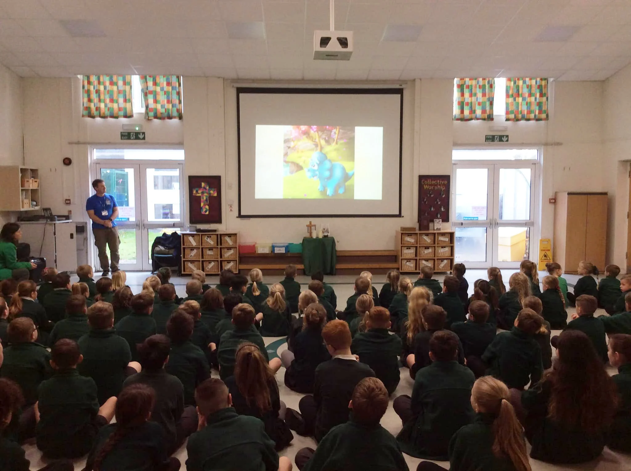 A large group of school children sitting on the floor attending a presentation in a school hall, with a man standing on the left side near a projector screen showing a picture of a blue dinosaur.