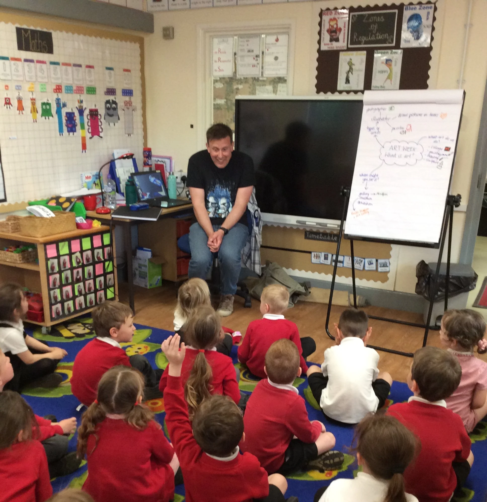 A man sits in front of a group of young children sitting on a colorful carpet in a classroom. The children are wearing red and white uniforms. The classroom has educational posters on the walls, a whiteboard with notes, and various classroom supplies.