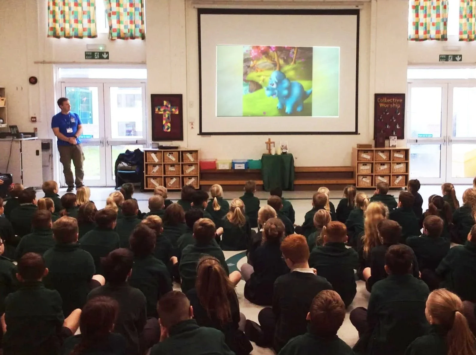A large group of children sitting on the floor in a school assembly hall, facing a screen with a colorful 3D animated elephant, while a man in a blue shirt stands to the left near the door.