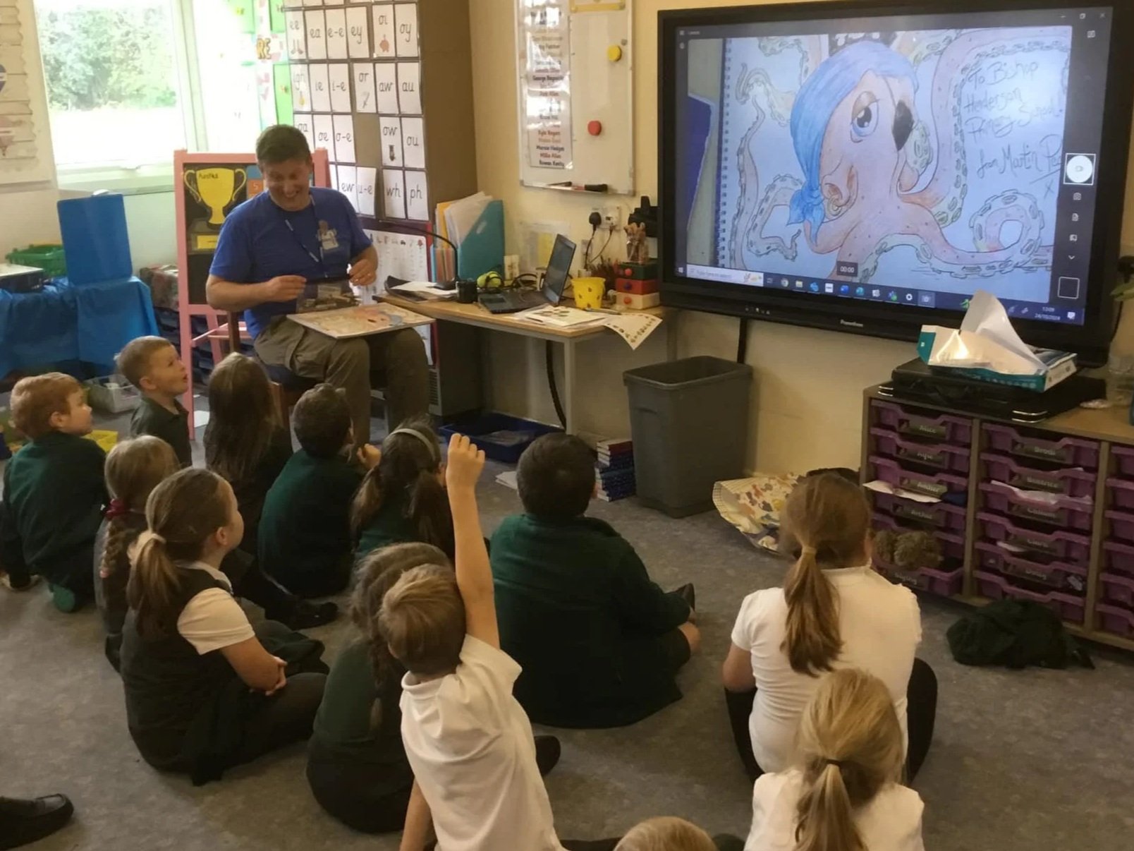 A teacher reading a story to a group of elementary school students in a classroom, with a large screen showing a digital drawing of an octopus wearing a blue hat.