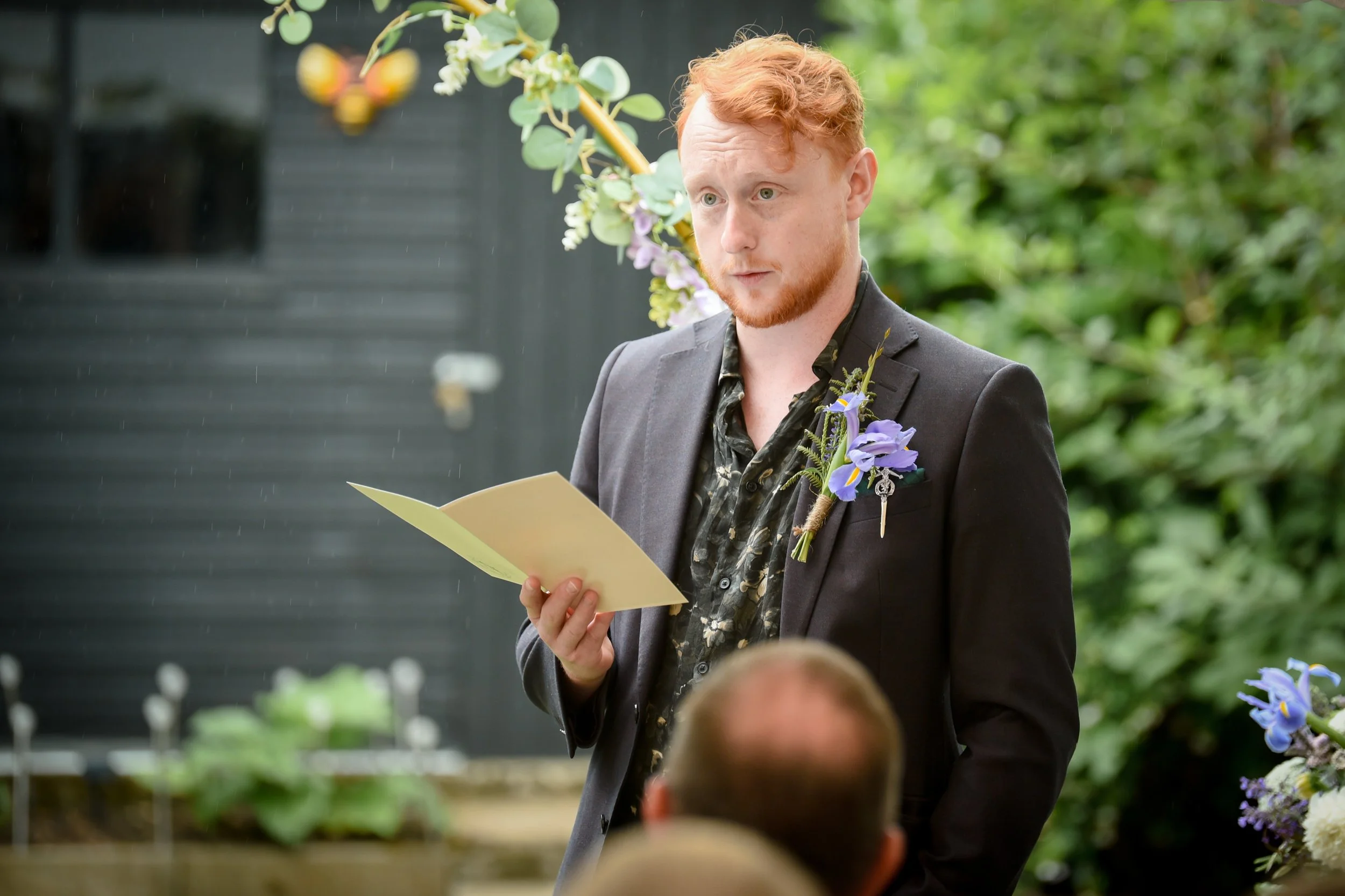 A man with red hair and a beard wearing a black suit and a flower boutonniere is reading from a piece of paper at an outdoor wedding. There are green plants and flowers in the background and a person with short hair seated in front of him.