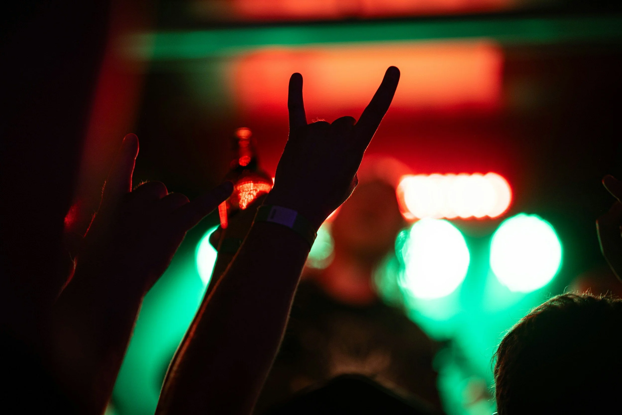 People raising their hands, with some making the 'rock on' gesture, at a concert or music event with colorful stage lights in the background.