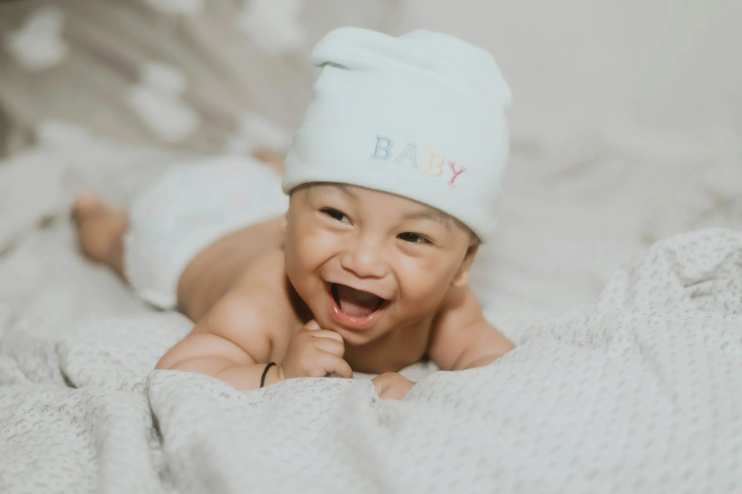 A small, smiling newborn baby lying on a soft white blanket, wearing a soft white hat with baby written on it