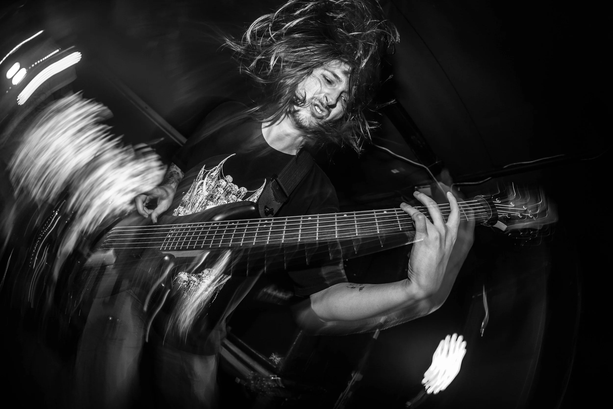 A musician with long hair playing an electric guitar passionately on stage in a dark setting, captured with motion blur in black and white.