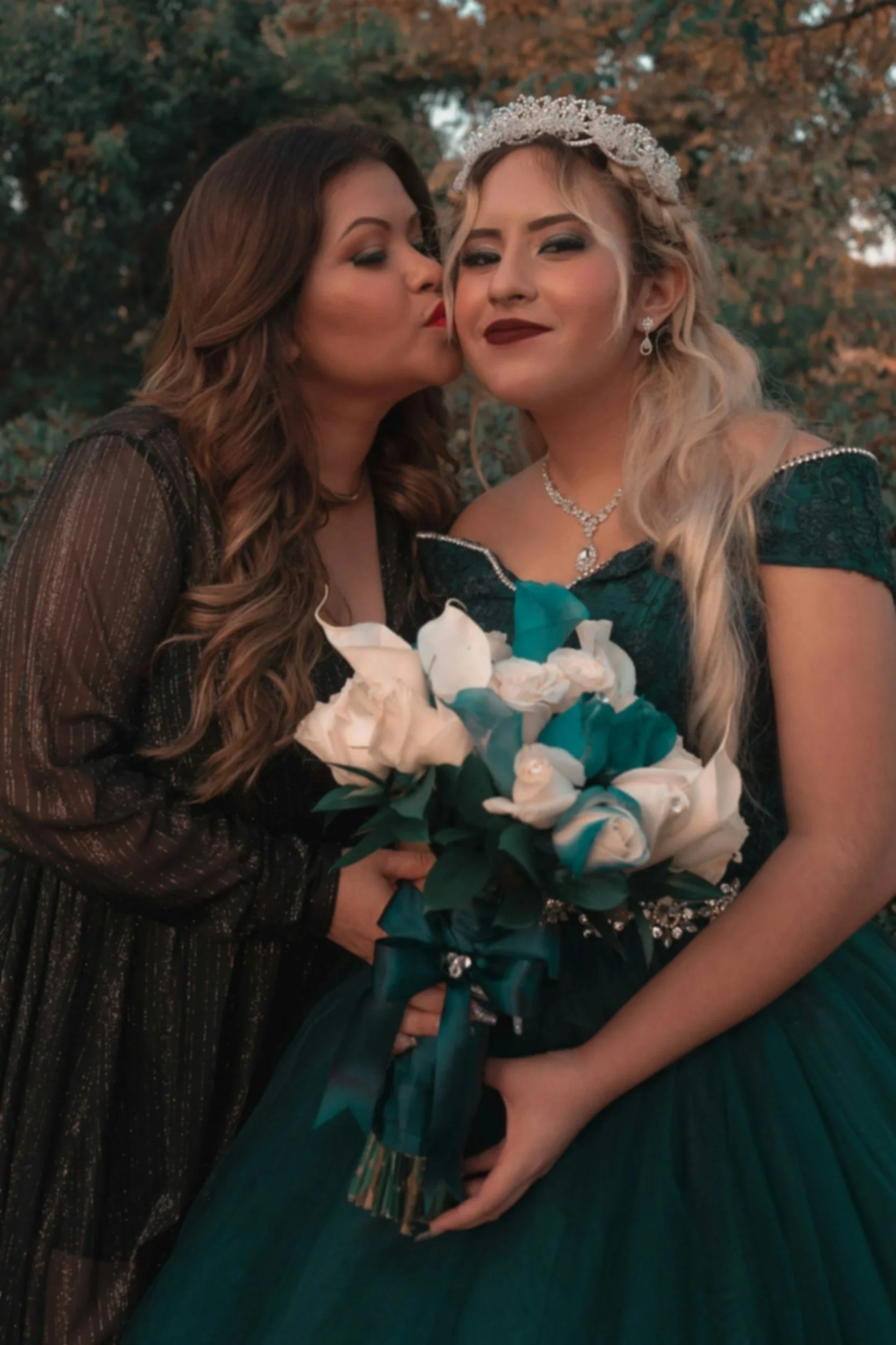 Two women in elegant wedding dresses, one wearing a tiara, holding a bouquet of white and teal flowers, outdoors with trees in the background, one woman is kissing the other on the cheek.