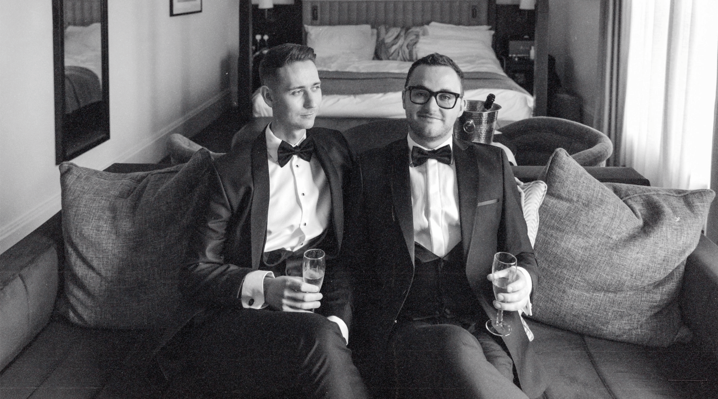 Two happy, stylish husbands to be holding tasteful champagne glasses, sat on a sofa, in an elegant hotel room.  