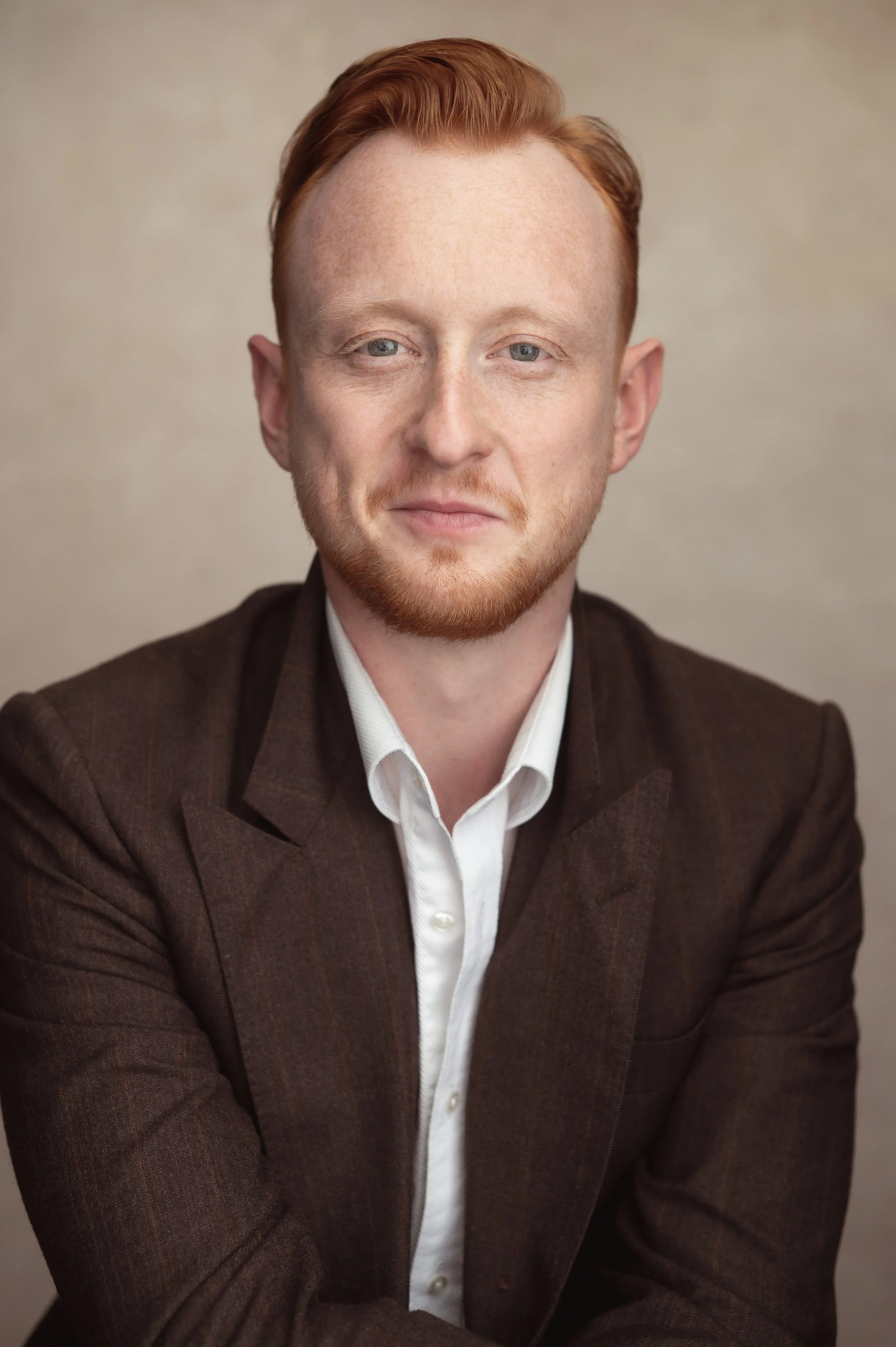A man with red hair and a light beard wearing a white shirt and dark brown blazer, smiling slightly, against a neutral background.