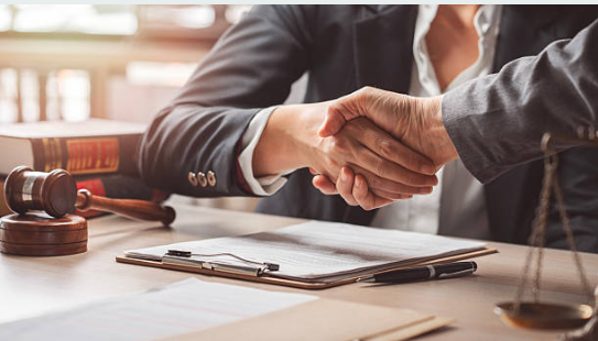 Close-up of two people shaking hands over a desk with legal documents, a gavel, and scales of justice in the background, indicating a legal or business agreement.