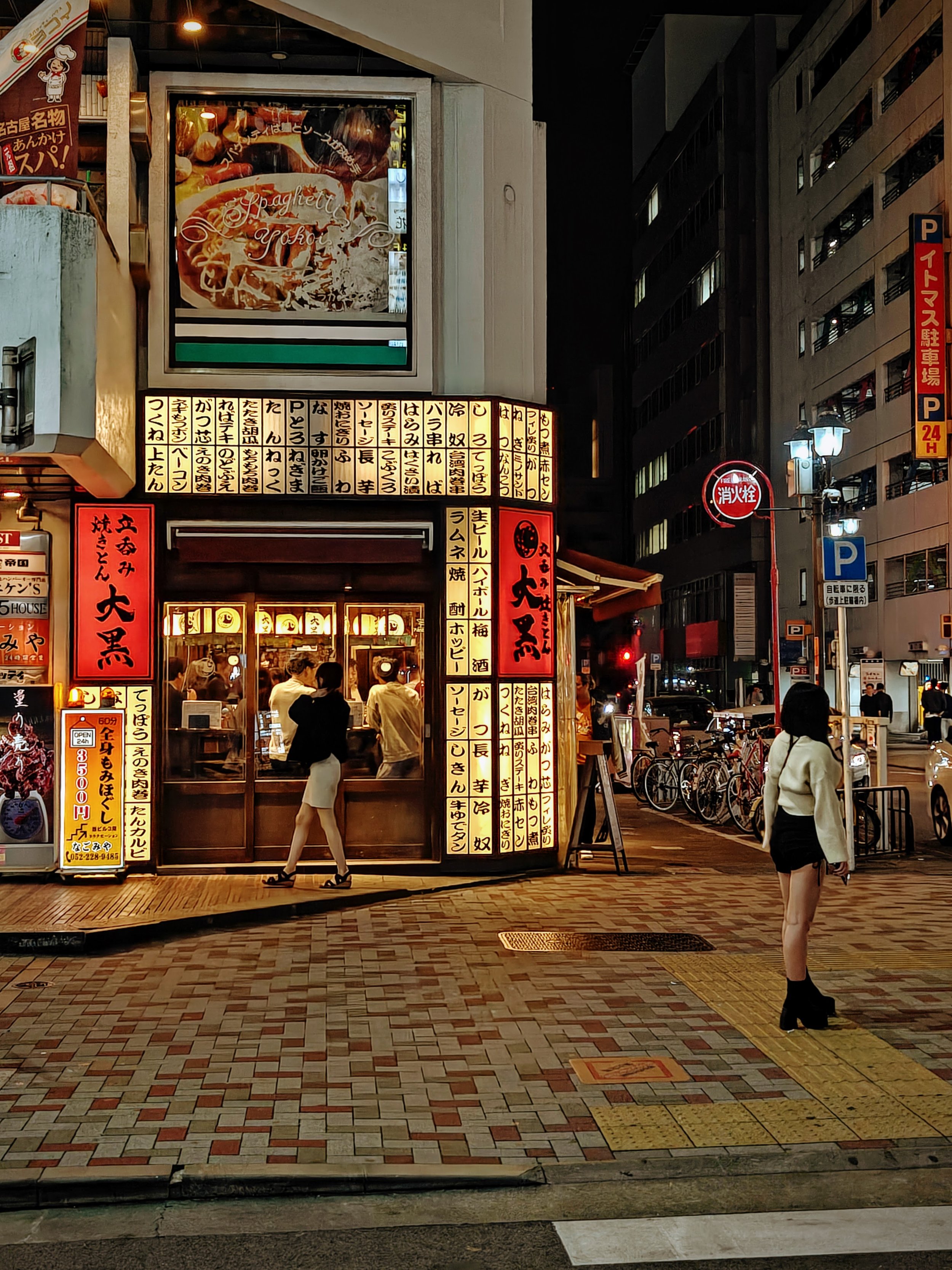 Night-time street scene outside Japanese restaurant with illuminated signs and people walking by, bicycles parked nearby.