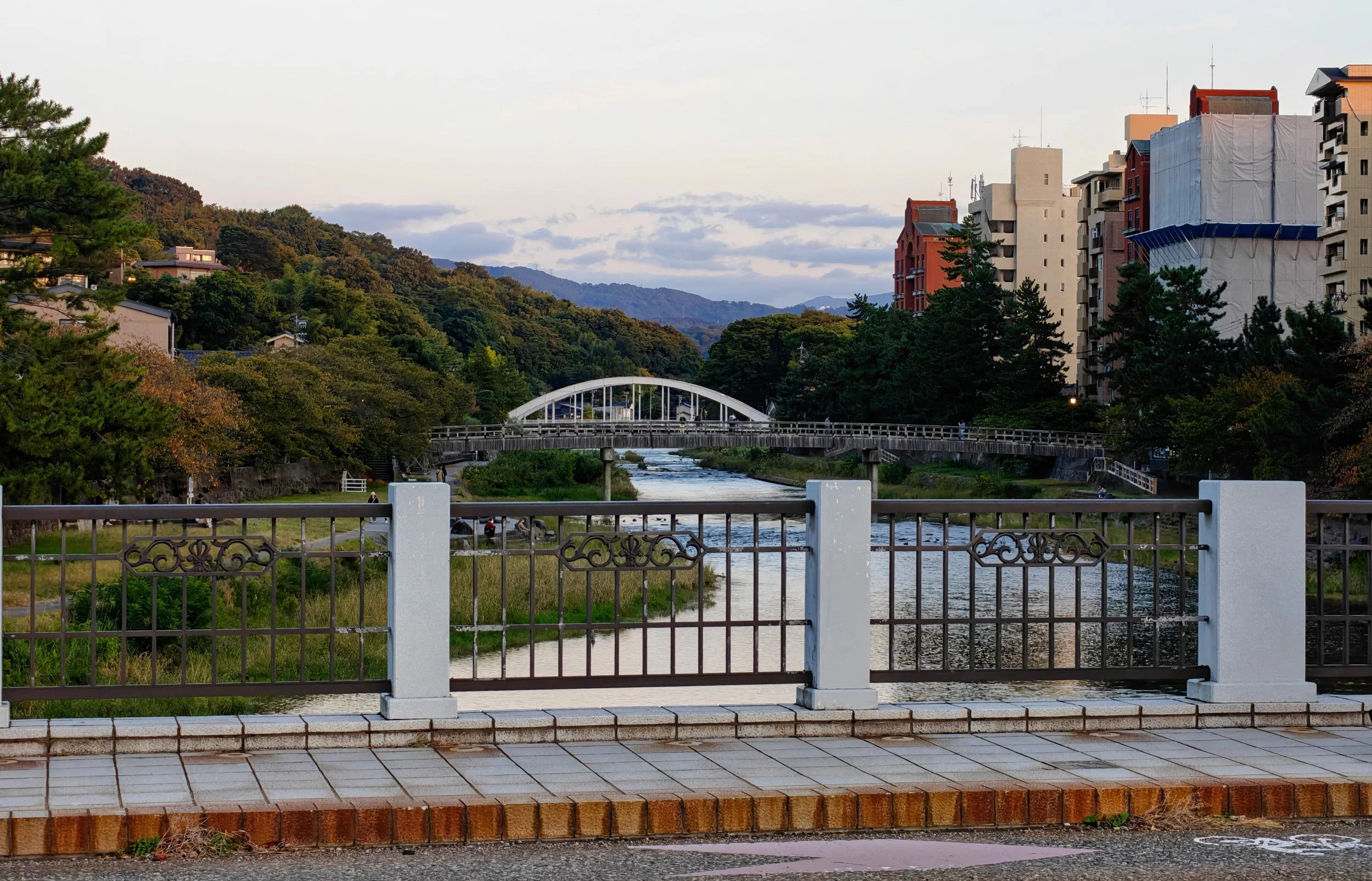 View of a river with bridges, surrounded by green trees and buildings, with mountains in the background, taken during late afternoon.