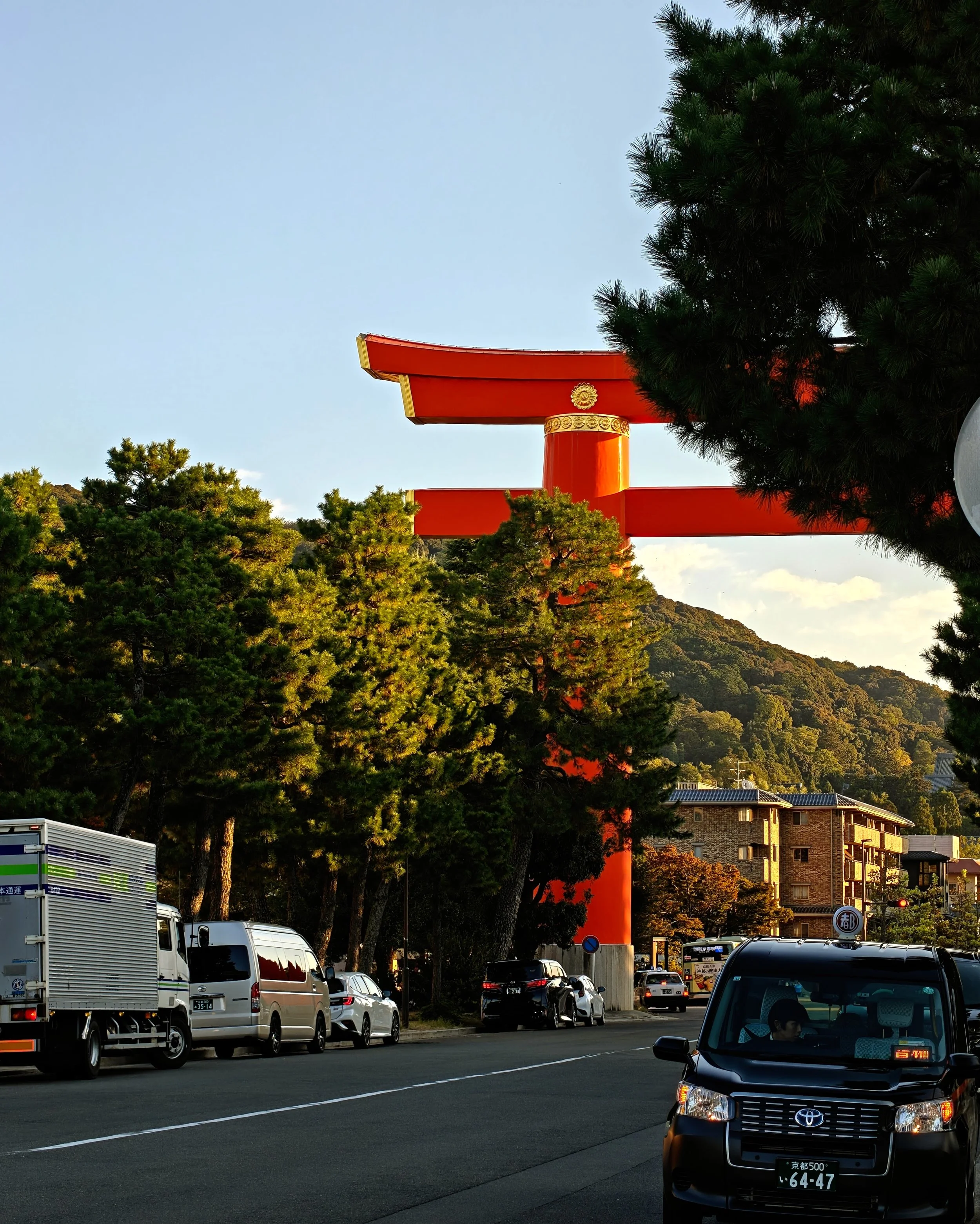 A large red torii gate partially obscured by trees, situated near a street with parked cars and modern buildings in the background, with a clear blue sky.