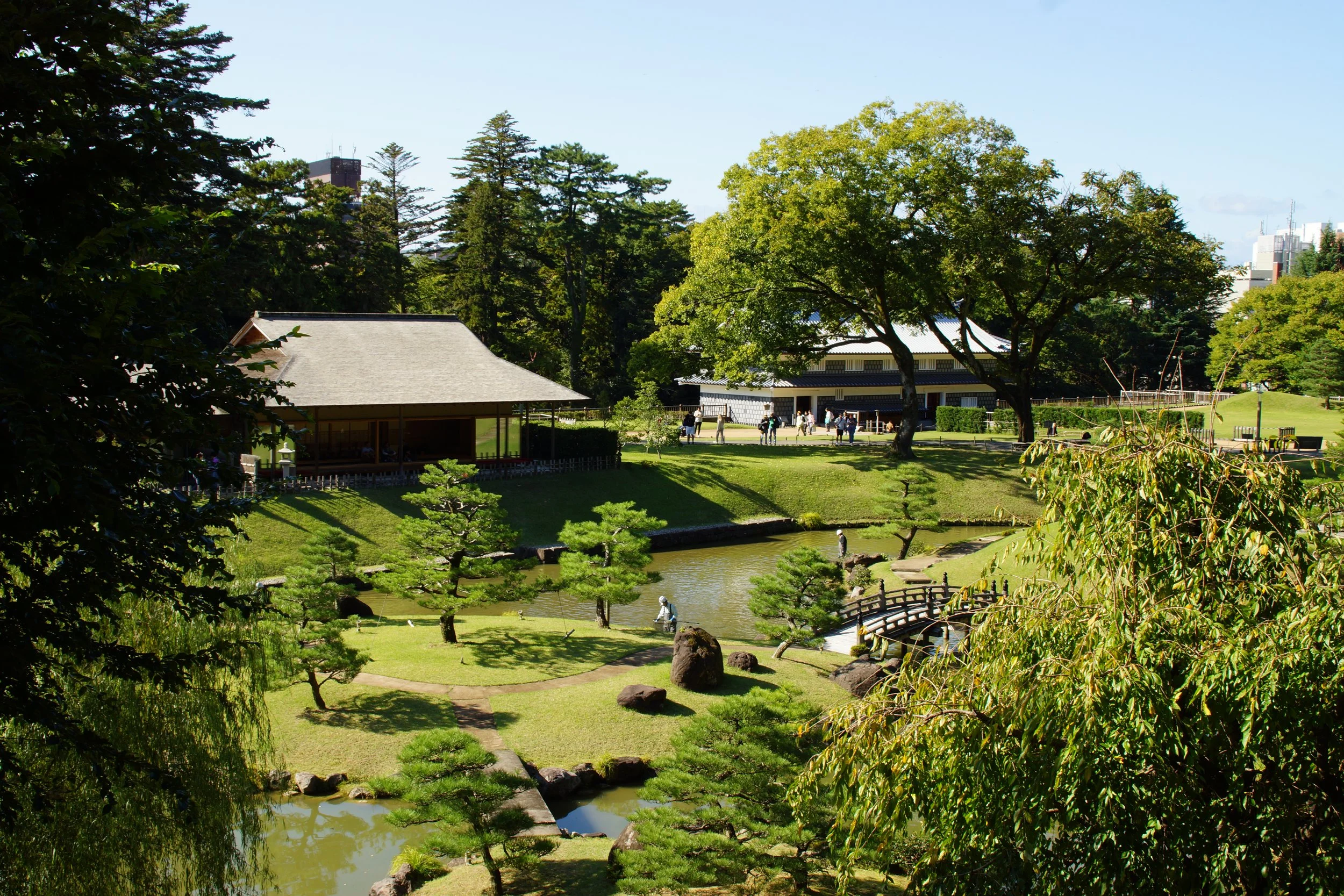 A peaceful Japanese garden featuring a pond, a small wooden bridge, manicured trees, and traditional buildings in the background, with visitors enjoying the scenery on a sunny day.