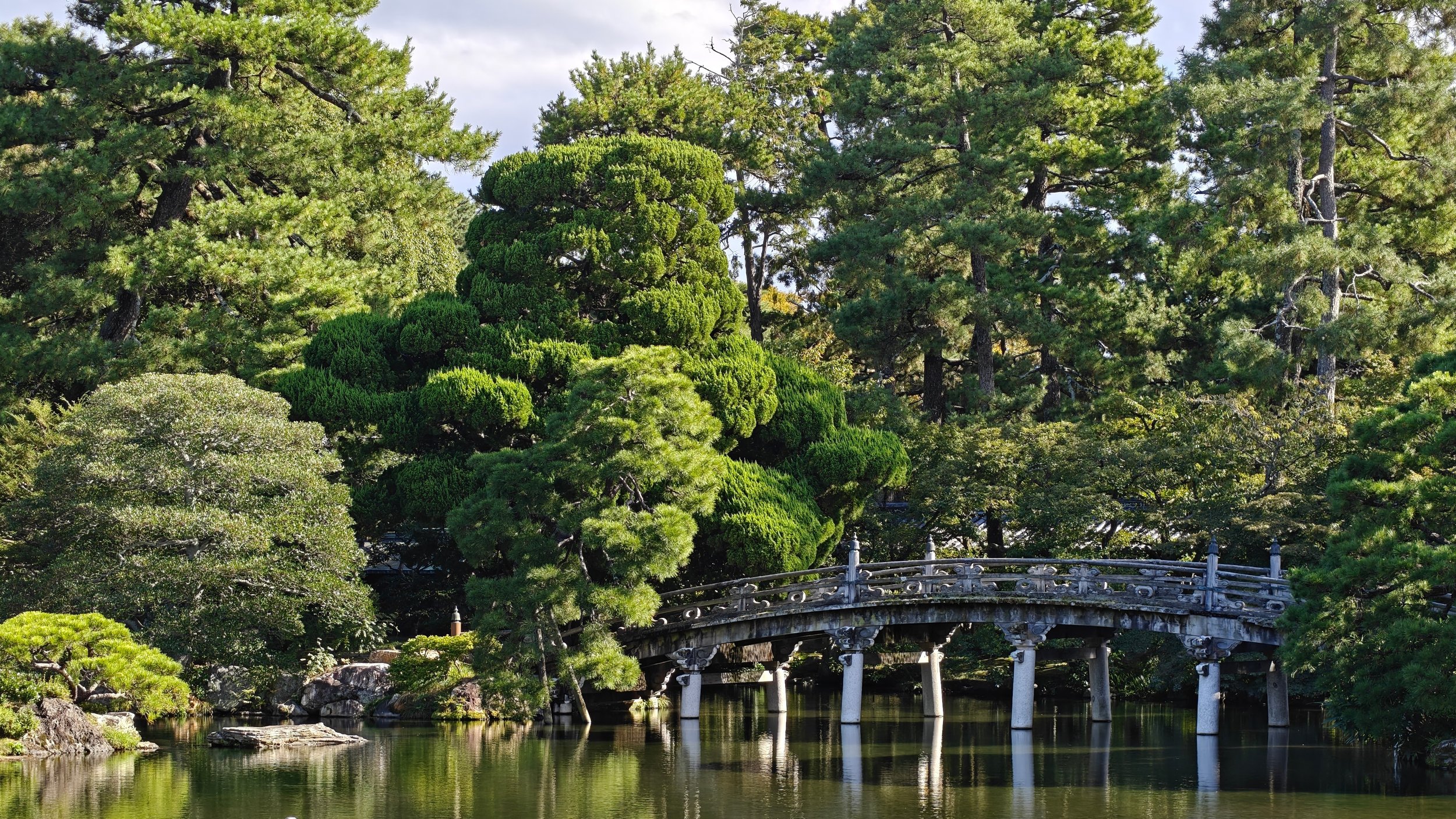 A traditional Japanese garden with a stone bridge over a pond, surrounded by lush green trees and shrubs.