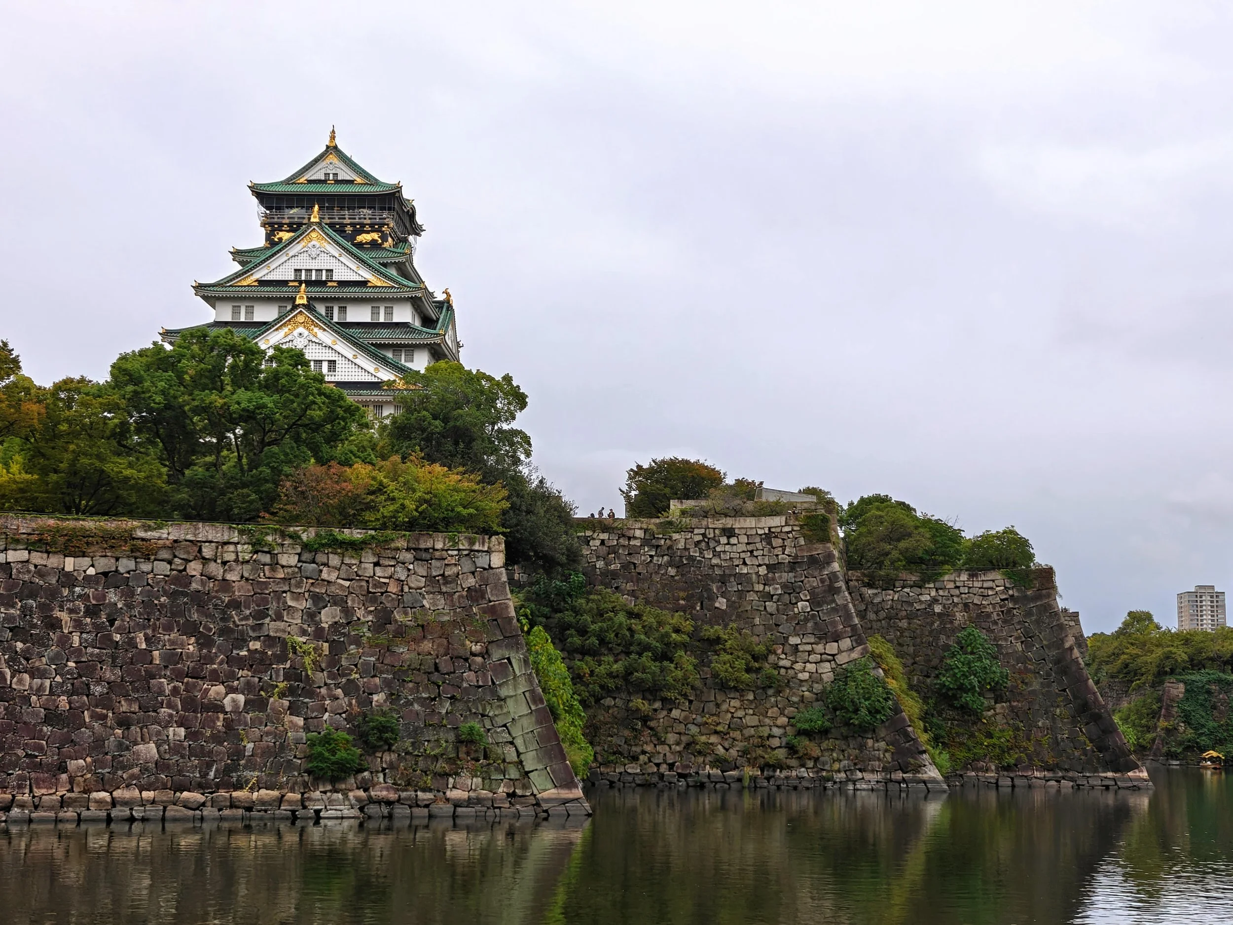 A traditional Japanese castle with multiple tiers and green roofs, situated on a stone fortress with a moat, surrounded by trees and an overcast sky.