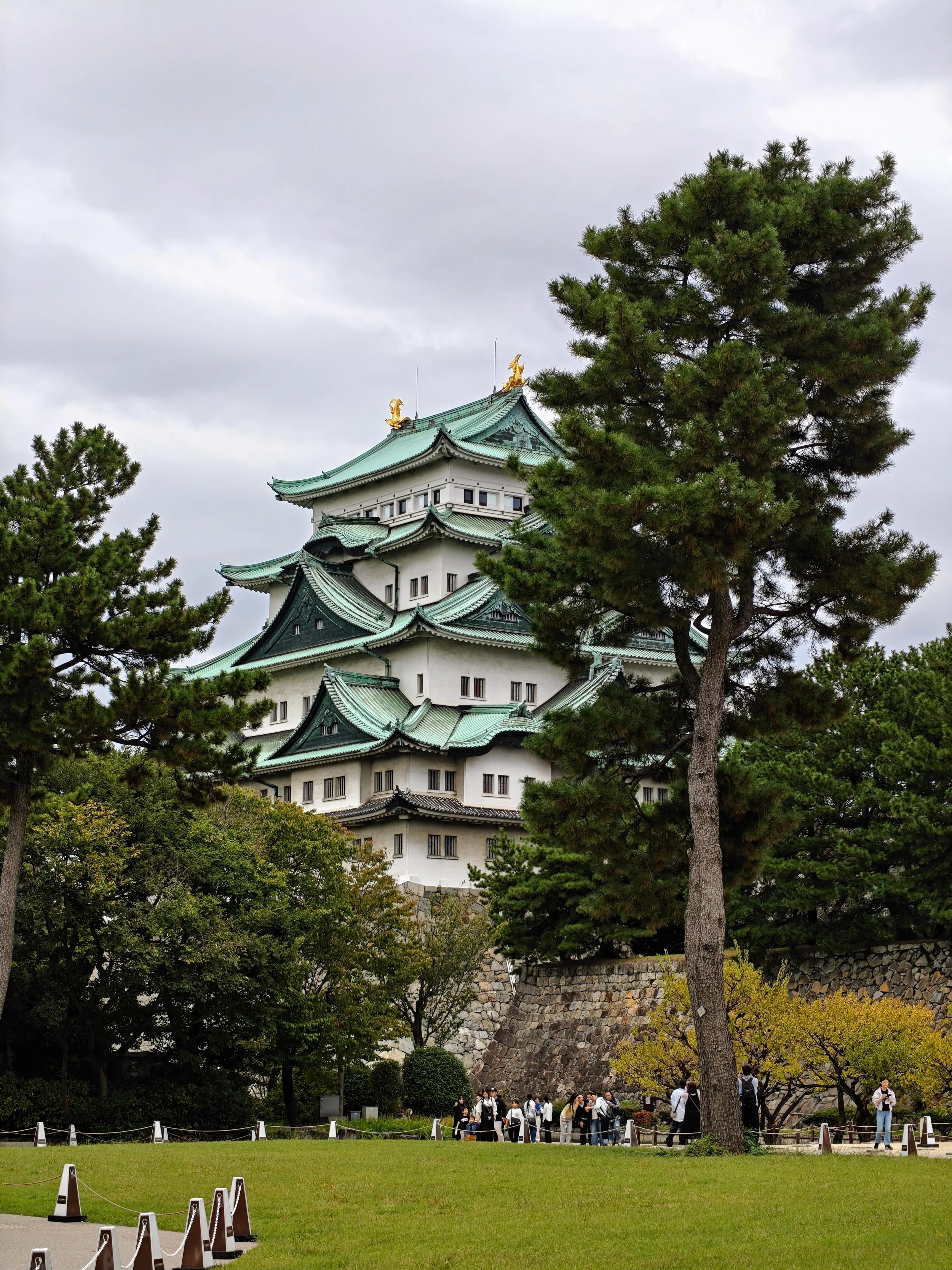 A traditional Japanese castle with multiple tiers and green roofs, surrounded by trees and a lawn, with a group of tourists nearby and a cloudy sky overhead.