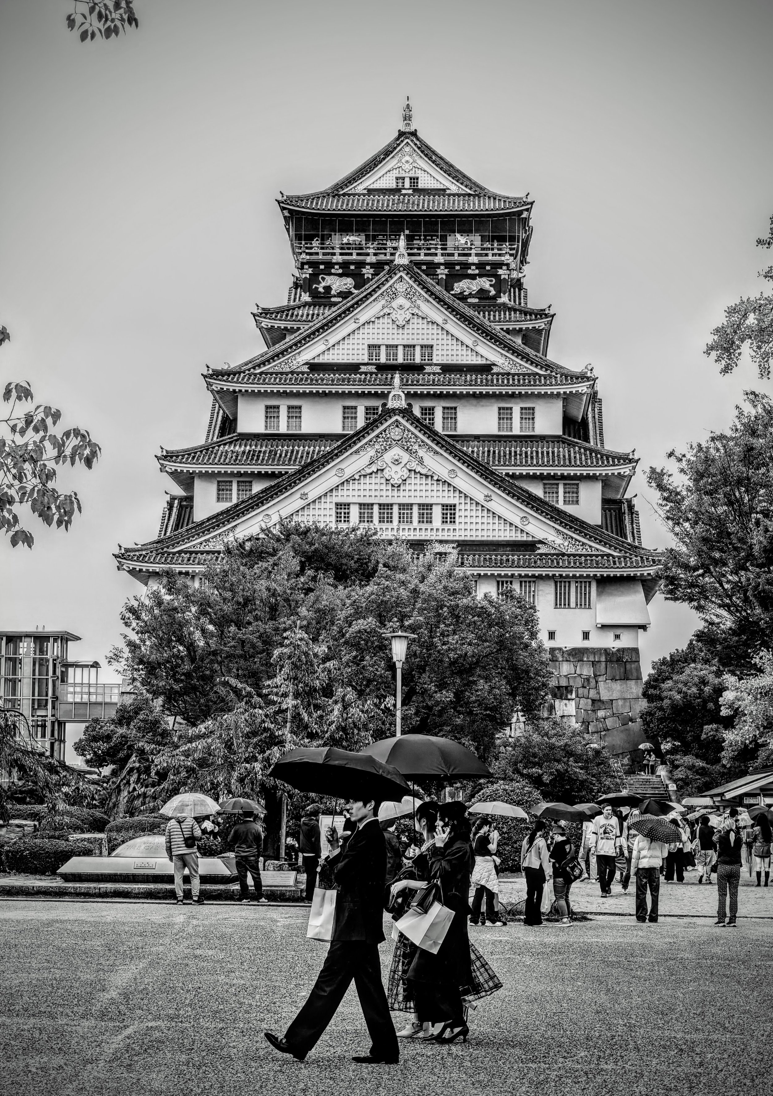 Black and white photo of a traditional Japanese castle with multiple tiers, surrounded by trees and a crowd of people holding umbrellas, suggesting it is raining.