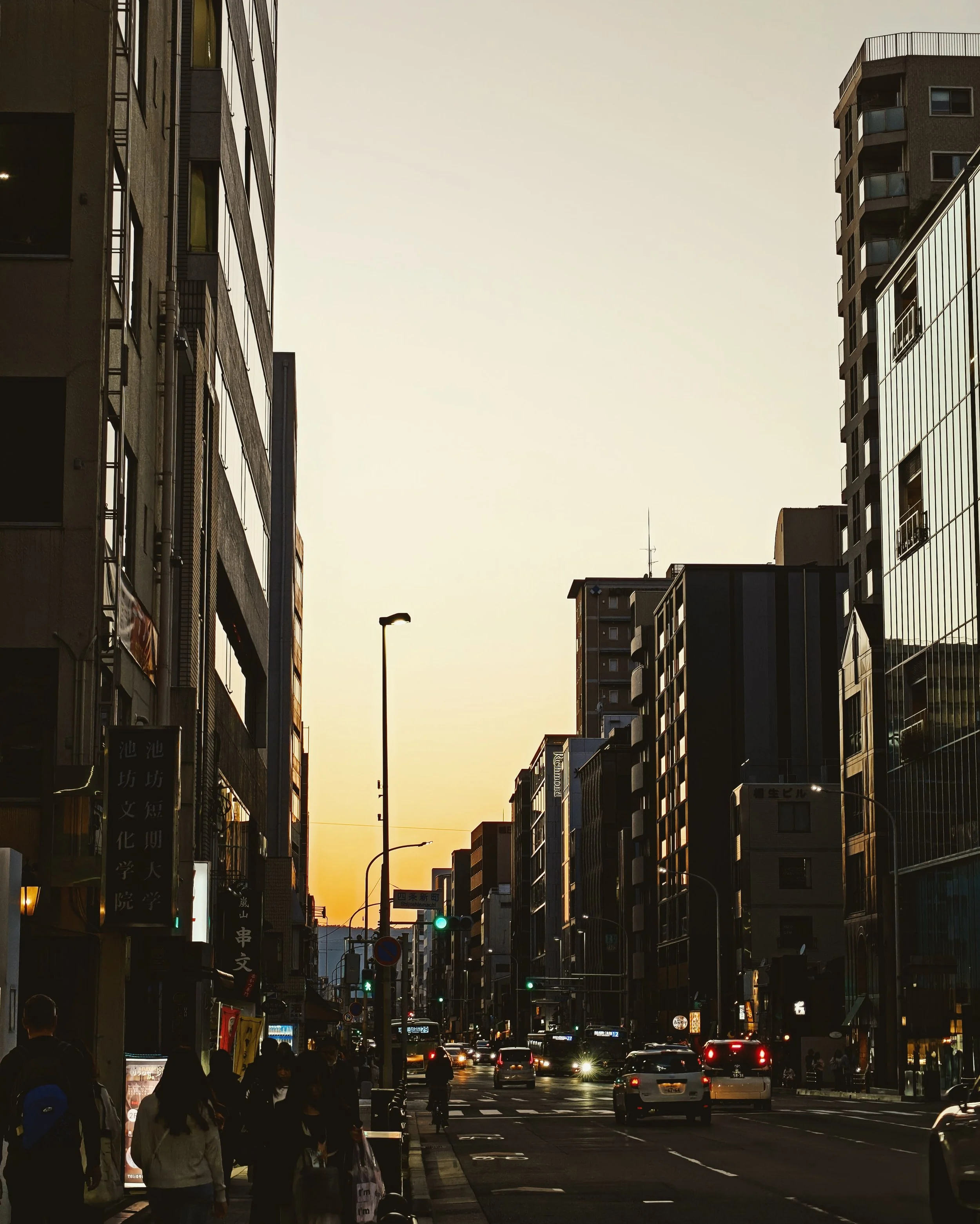 City street with tall buildings on both sides, evening sunset, cars and pedestrians.