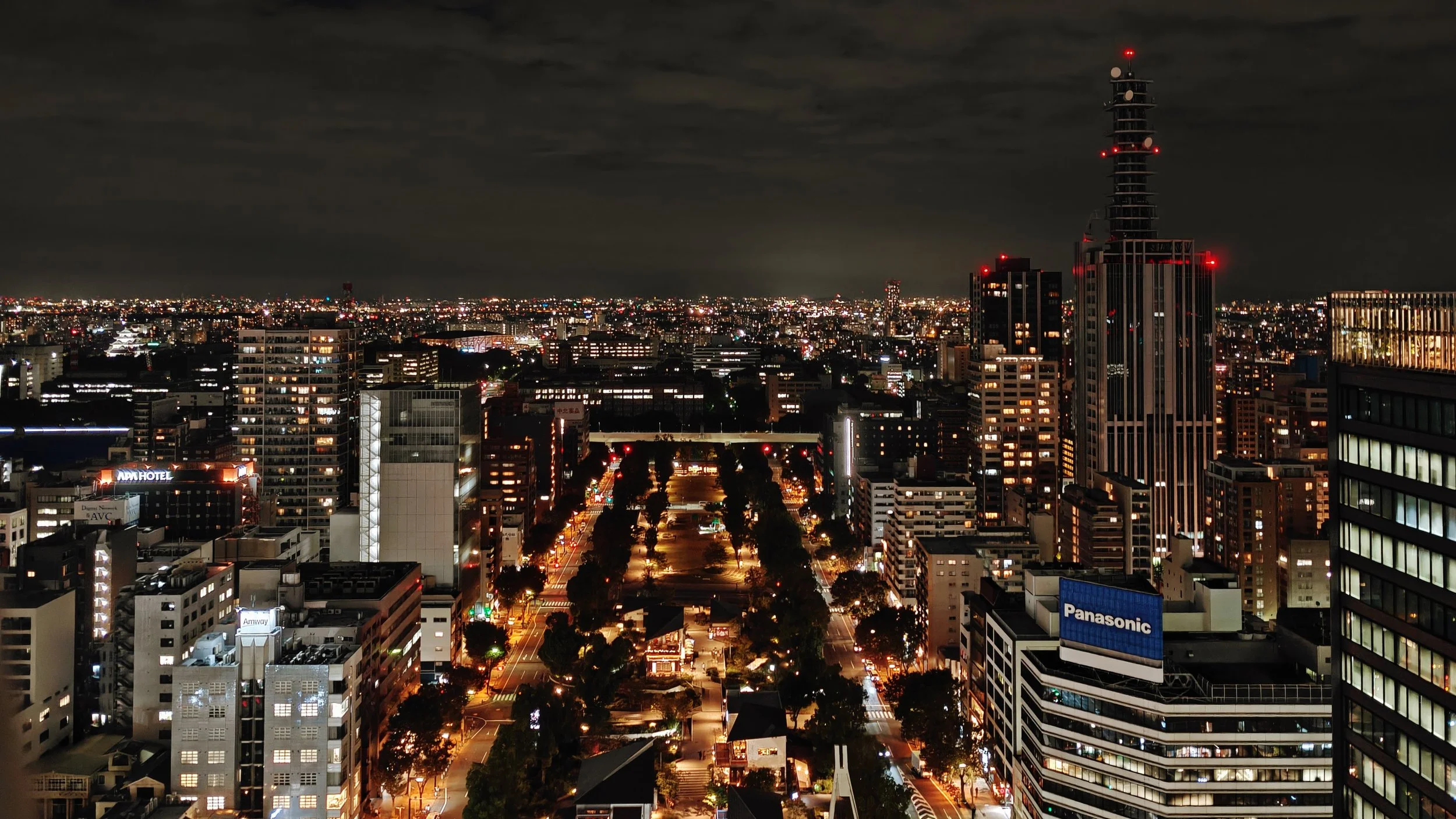 Nighttime cityscape with tall buildings illuminated, a wide avenue lined with streetlights, trees, and smaller buildings.