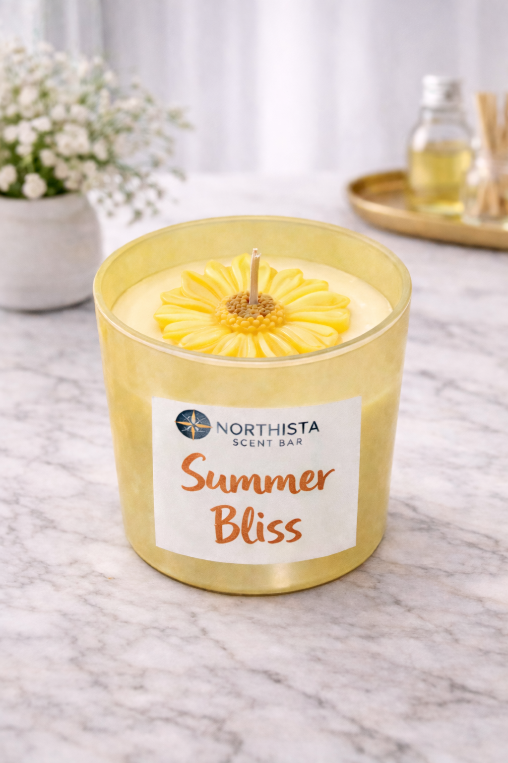A yellow candle with a sunflower design on top, labeled 'Summertime Bliss' from Northista Scent Bar, placed on a marble surface with a background of a potted white flower and bottles of oil and reed diffuser.