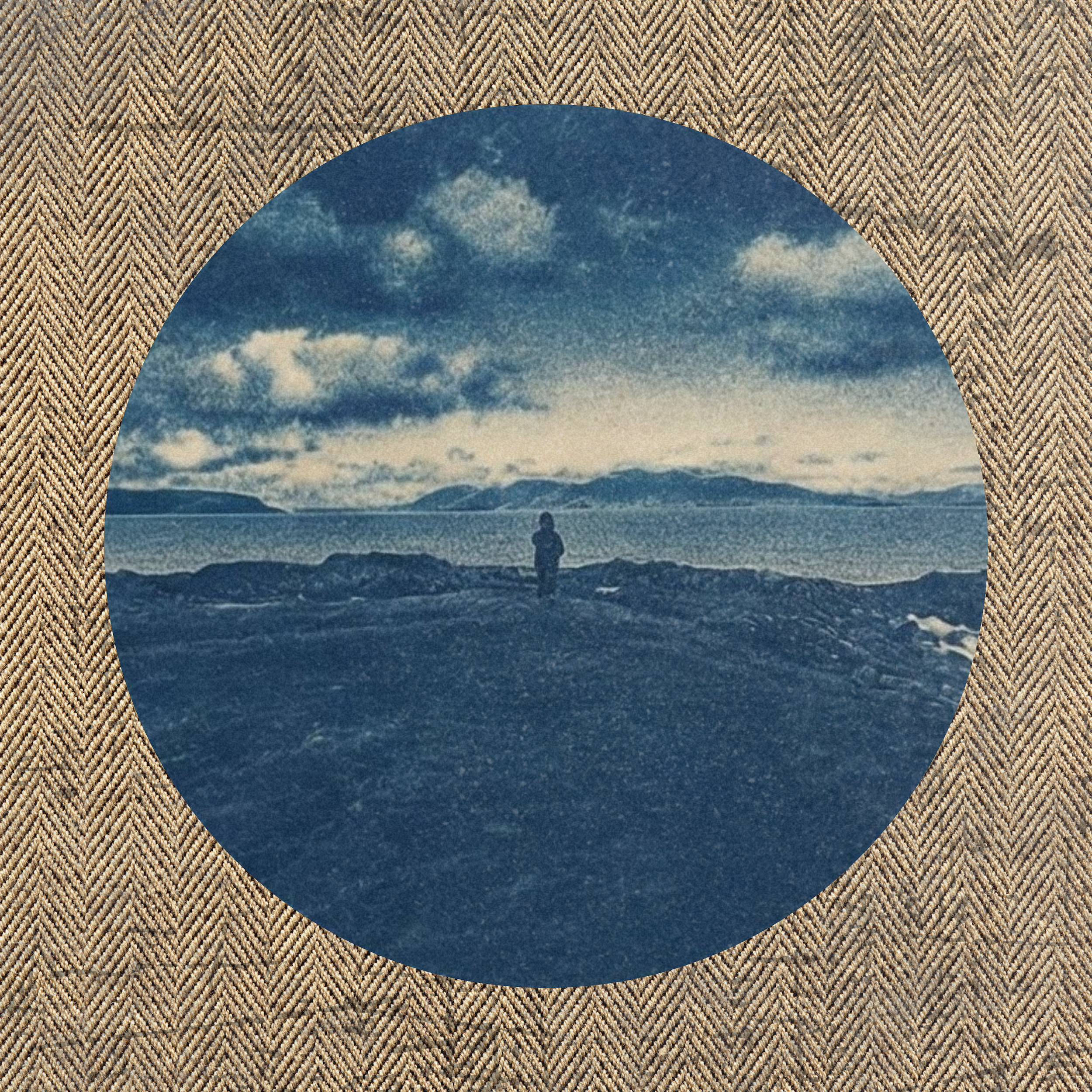 A person standing on rocky terrain near water with mountains and a cloudy sky in the background.