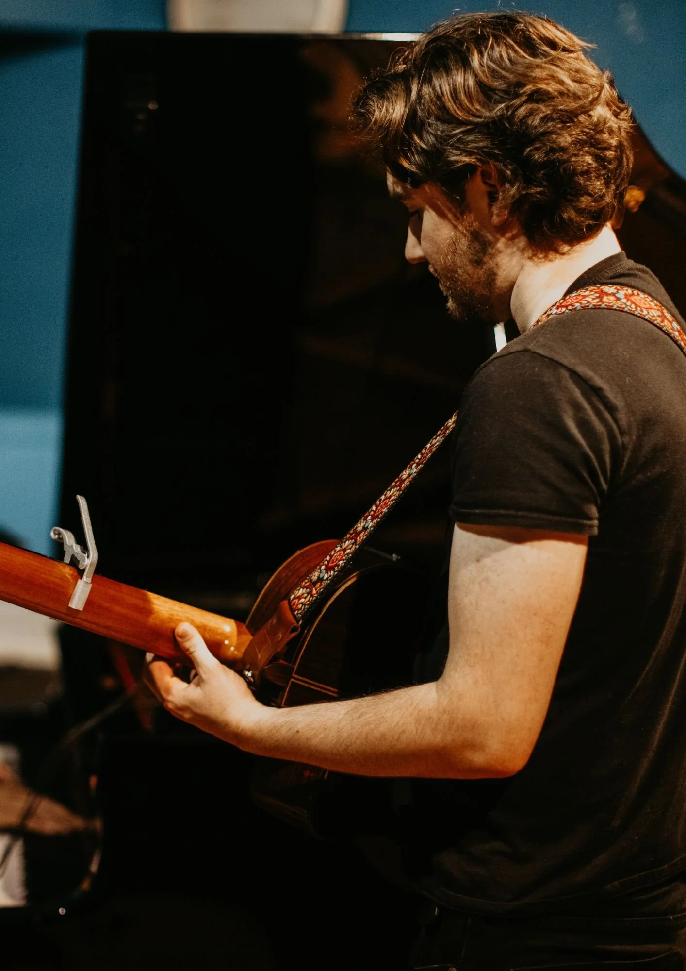 A man with curly brown hair, wearing a black t-shirt, playing an acoustic guitar with a colorful woven strap in a dimly lit room.