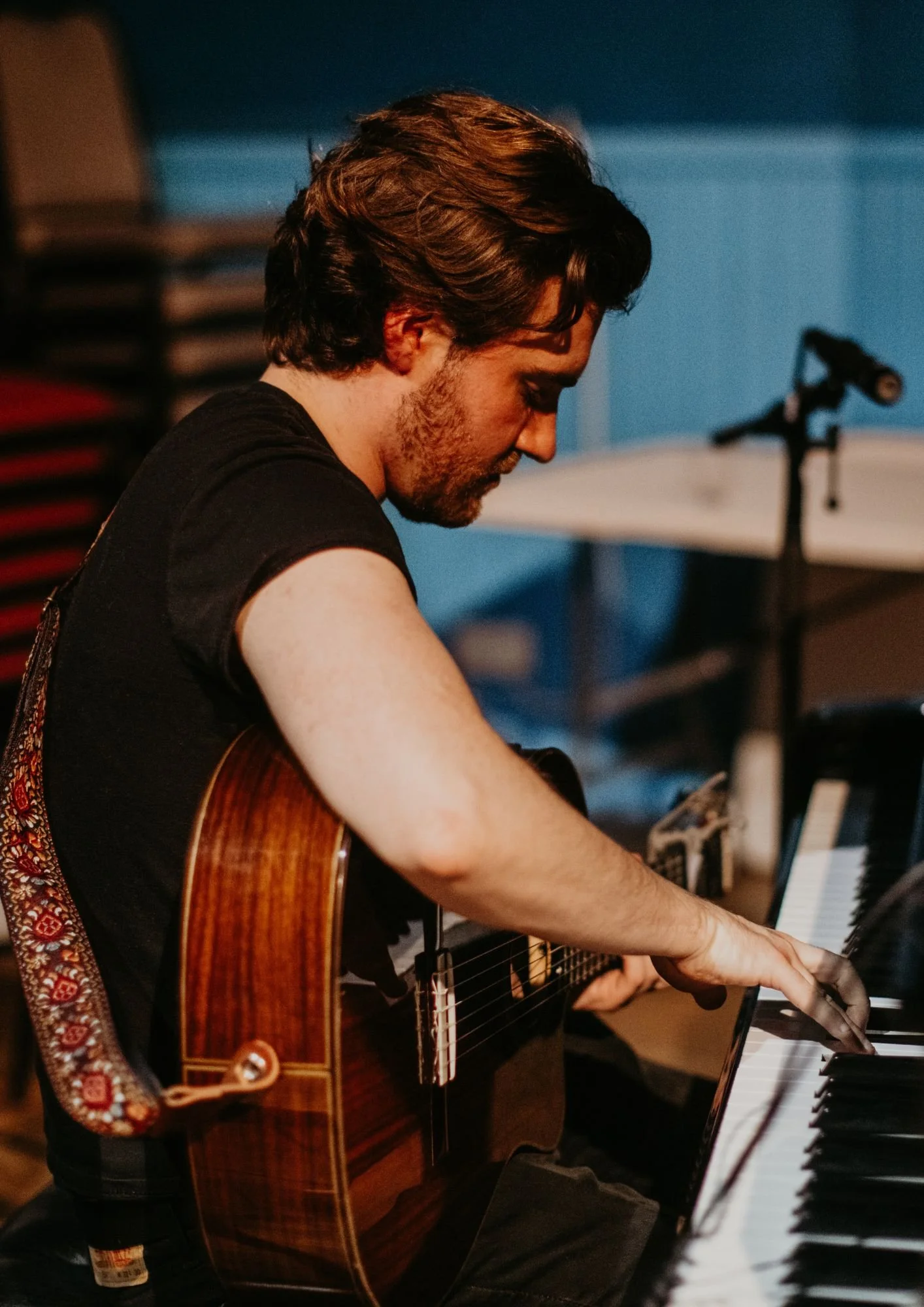 A man with dark hair and beard playing an acoustic guitar and keyboard in a recording studio.