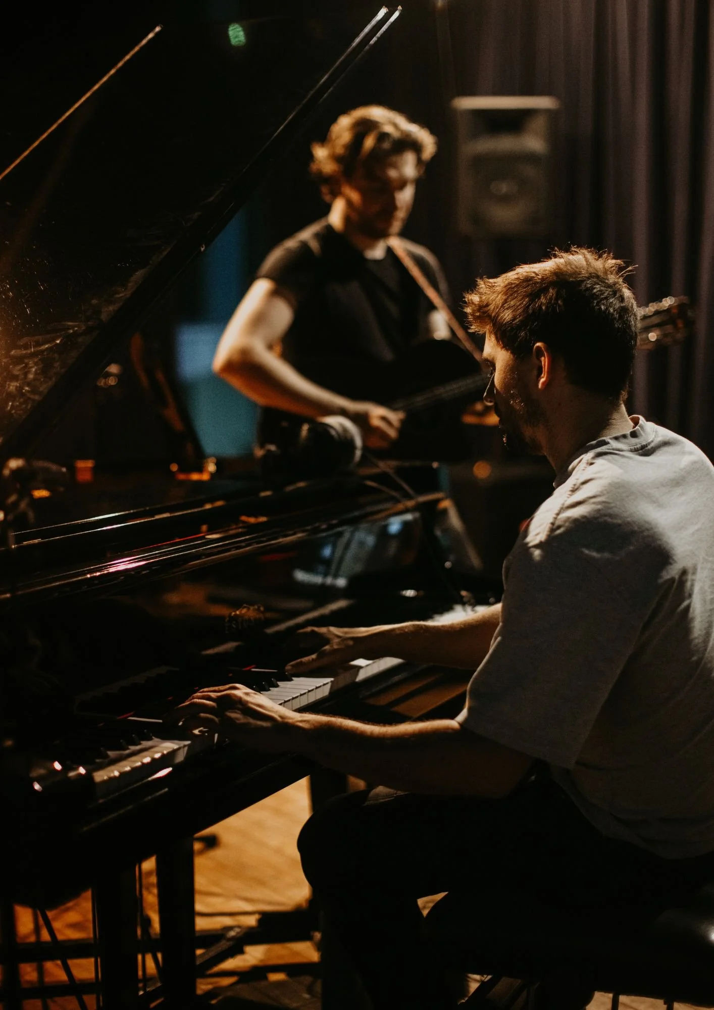 Two musicians performing in a dimly lit room: a man playing the piano and a woman playing the guitar.