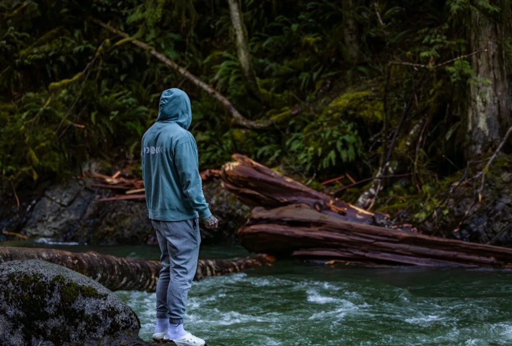 Person in a blue hoodie and gray pants standing on rocks by a river in a forest.