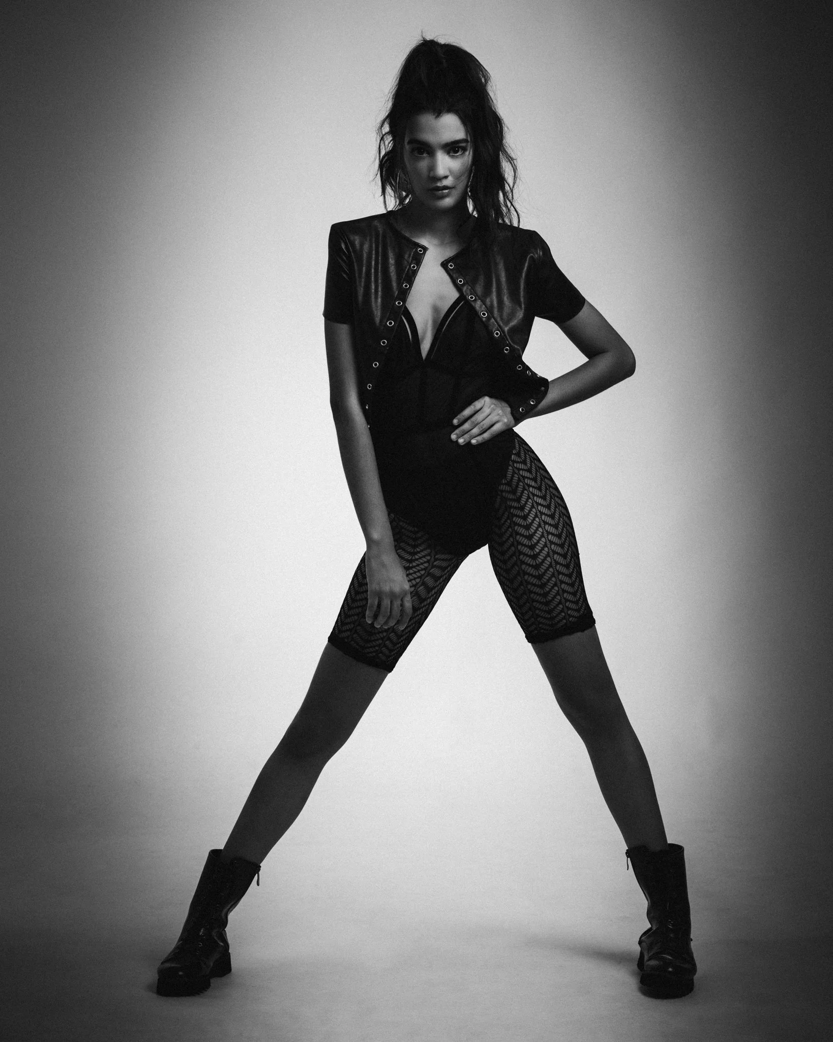A woman dressed in black fashion poses with legs apart, wearing a leather jacket, lace bodysuit, patterned tights, and combat boots in a studio setting.