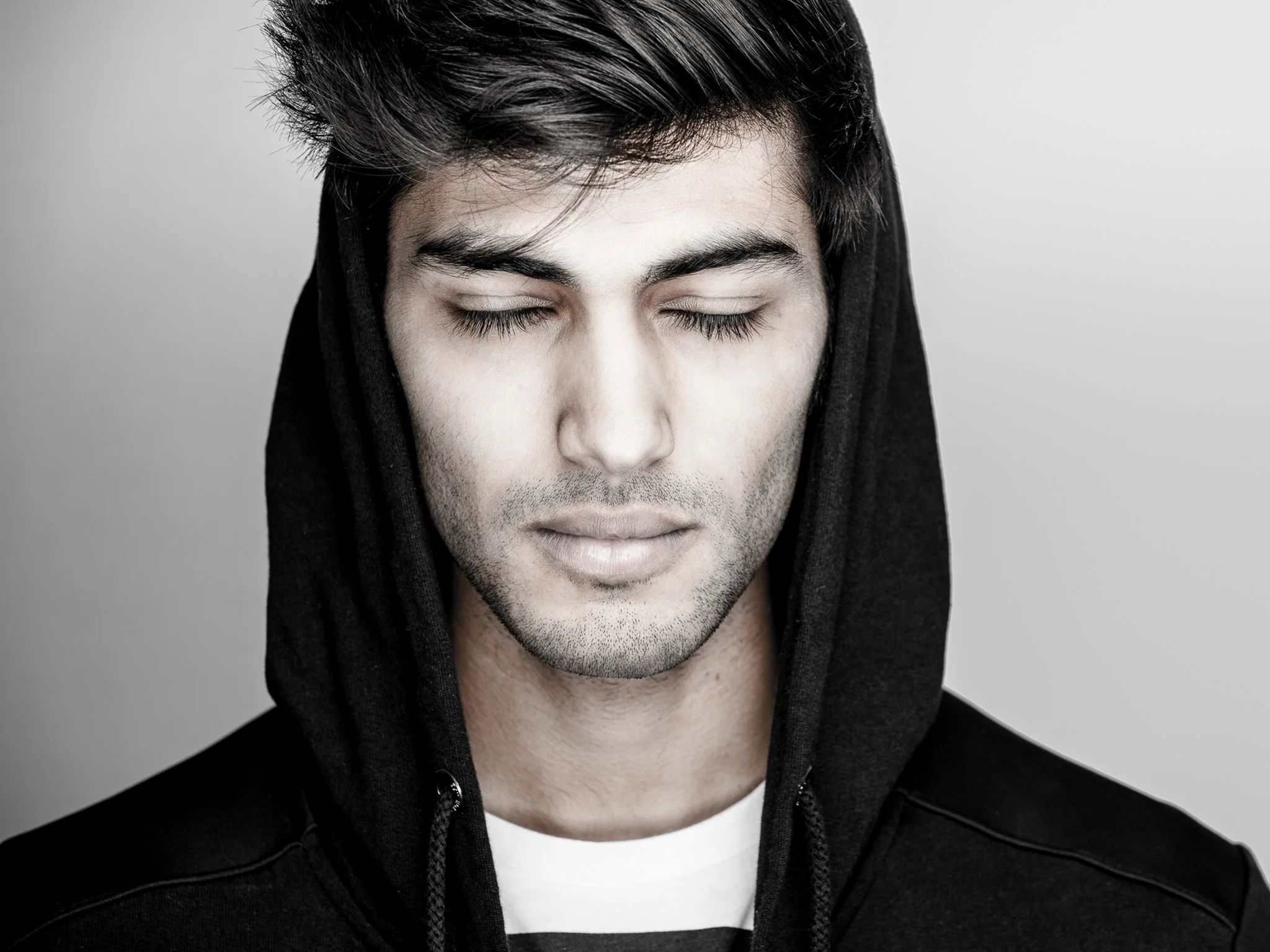 A young man with closed eyes, dark hair, and light skin, wearing a black hoodie and a white shirt, against a neutral background.