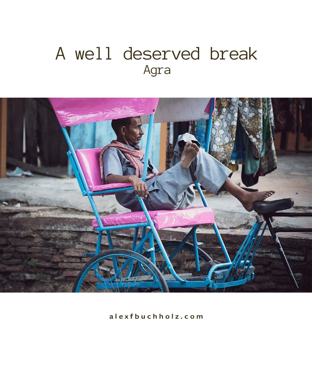 A man sitting in a pink rickshaw on a street, wearing a gray outfit and scarf, with his feet resting on the rickshaw's support, surrounded by hanging clothing.