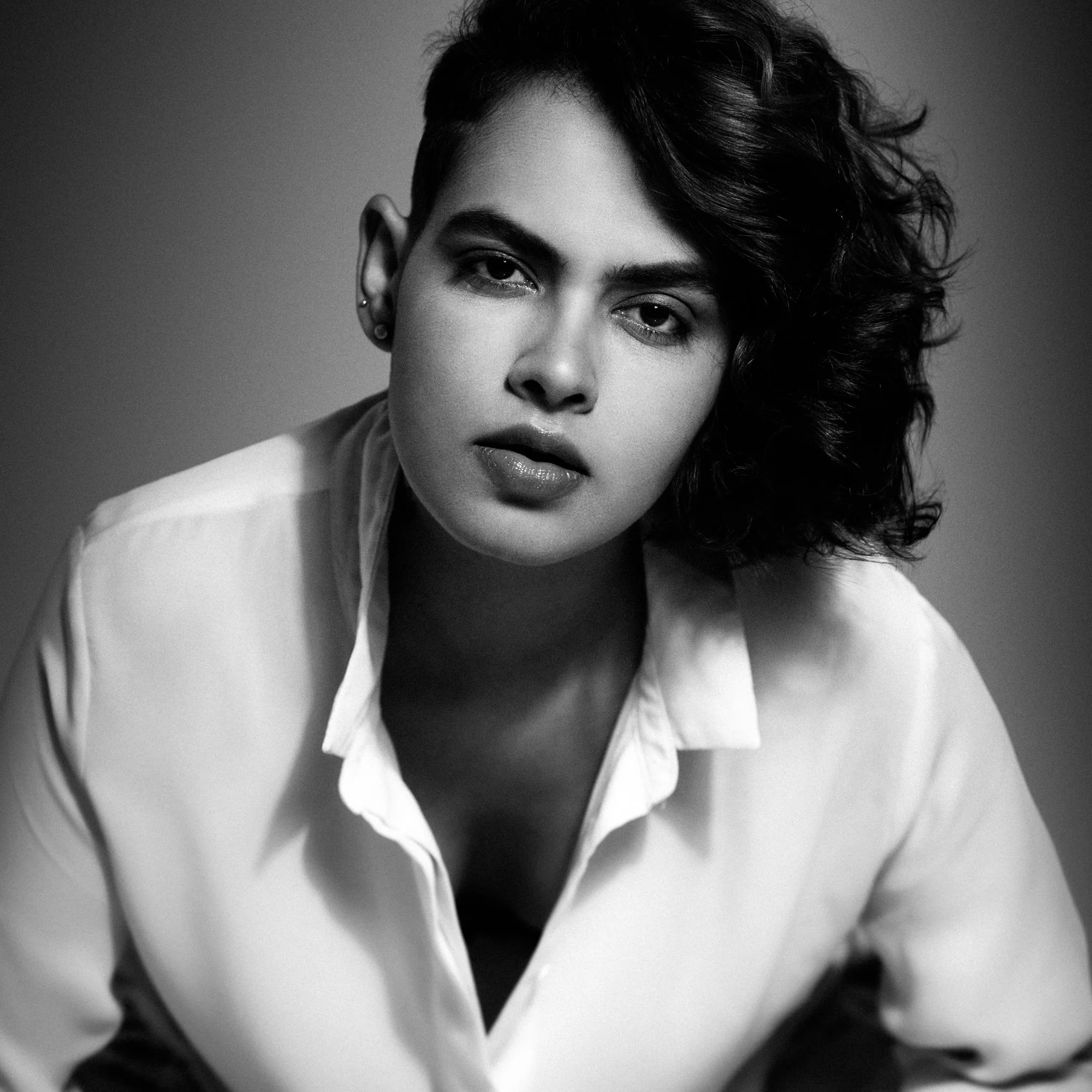 Black-and-white portrait of a woman with dark, wavy hair, wearing a white shirt, looking directly at the camera with a serious expression.