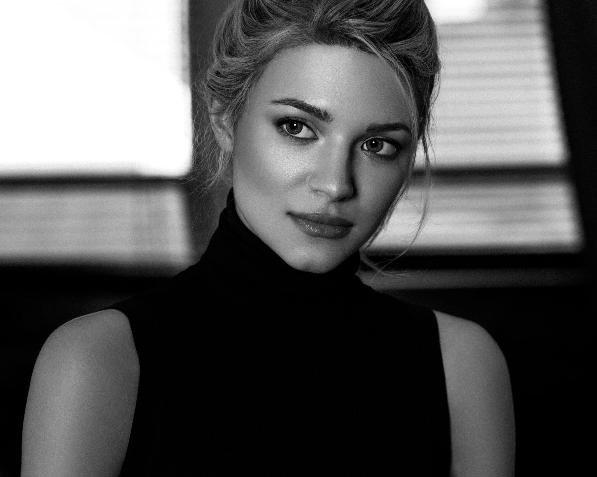 A black-and-white portrait of a young woman with blonde hair, wearing a sleeveless black turtleneck top, looking at the camera with a neutral expression.