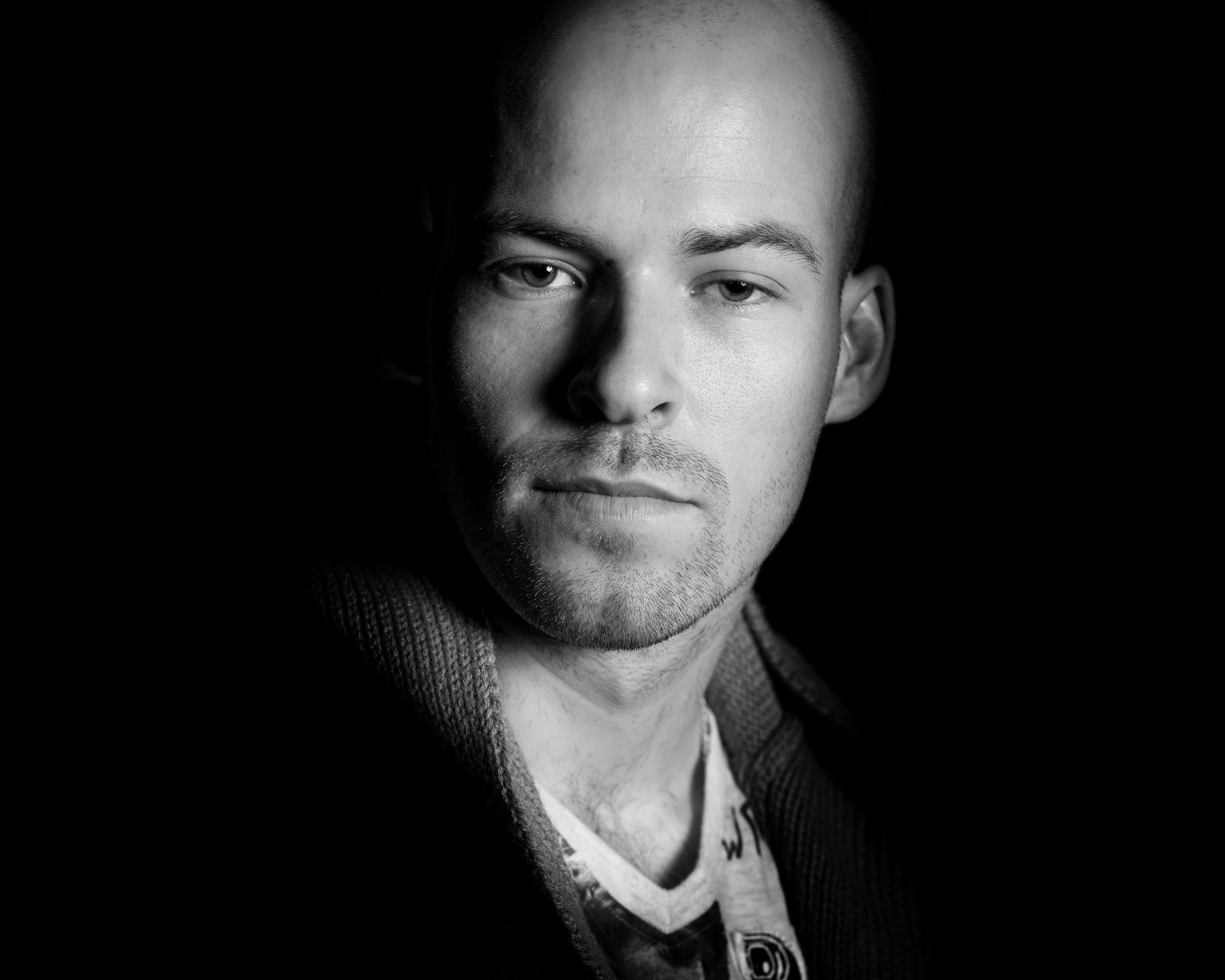 A black and white portrait of a young man with a serious expression, looking slightly to the right of the camera, with lighting highlighting one side of his face.