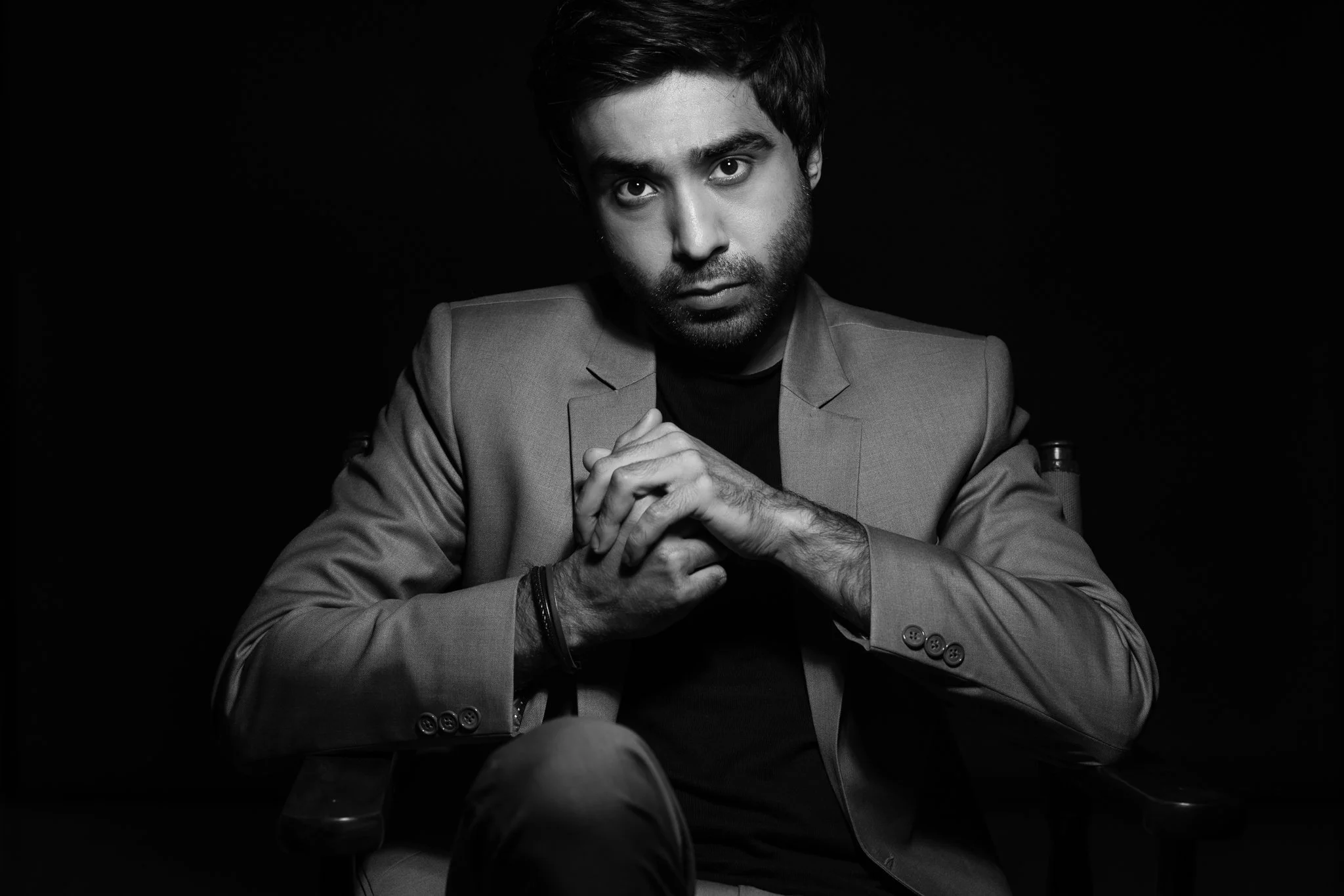 Black and white portrait of a young man with dark hair and beard, wearing a suit jacket and black shirt, sitting against a black background, gazing directly at the camera with a serious expression.