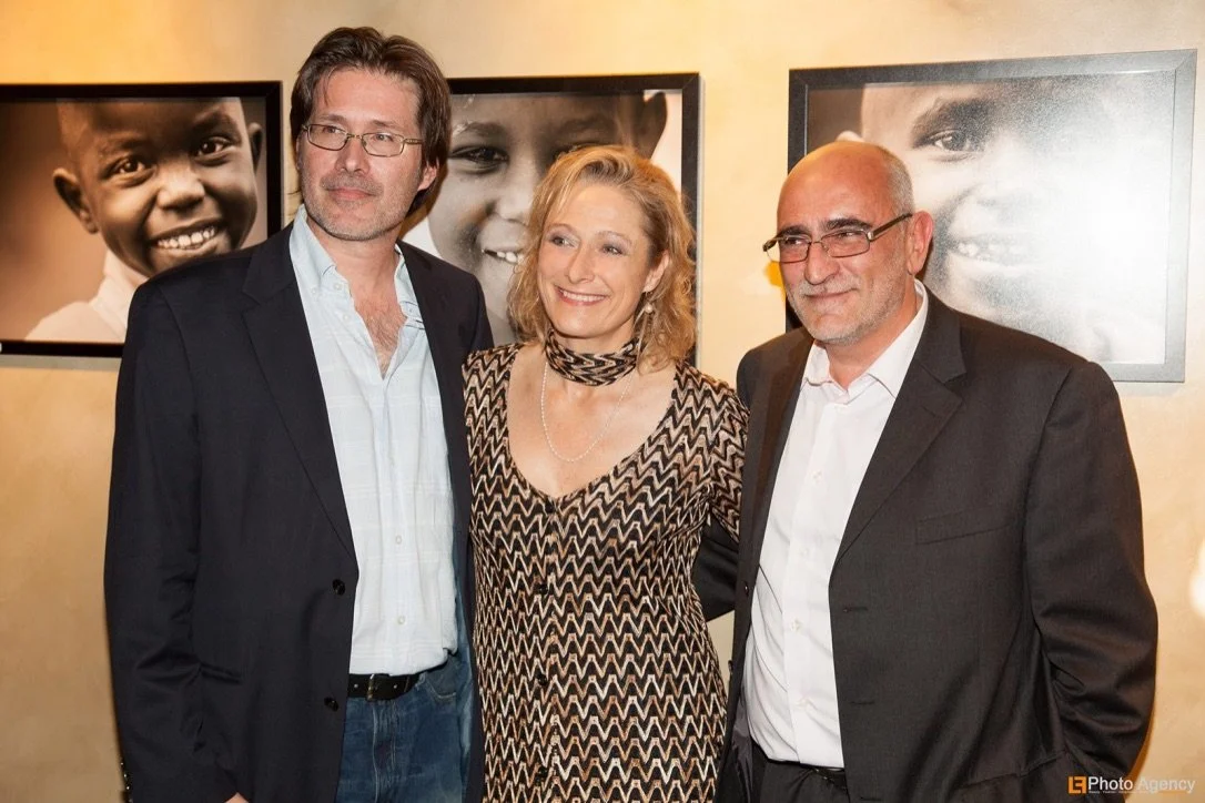 Caroline Goodall, Pere Larregual and Alex F Buchholz standing side by side at an event, smiling, with large framed black-and-white photos of children in the background. Image taken during the Care Highway fundraising exhibition.
