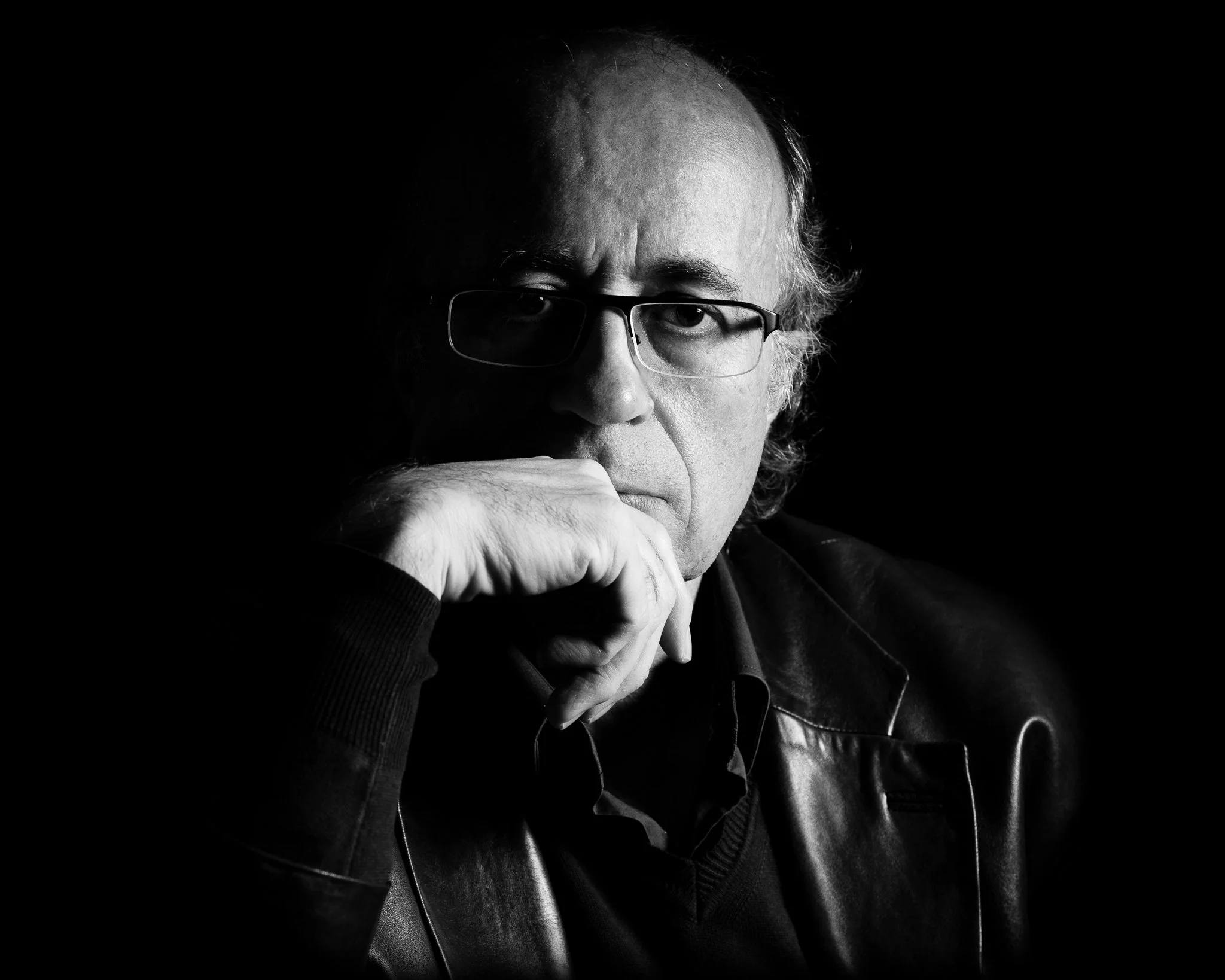 Black and white portrait of a man wearing glasses and a leather jacket, with his hand resting on his chin, looking contemplative against a dark background.