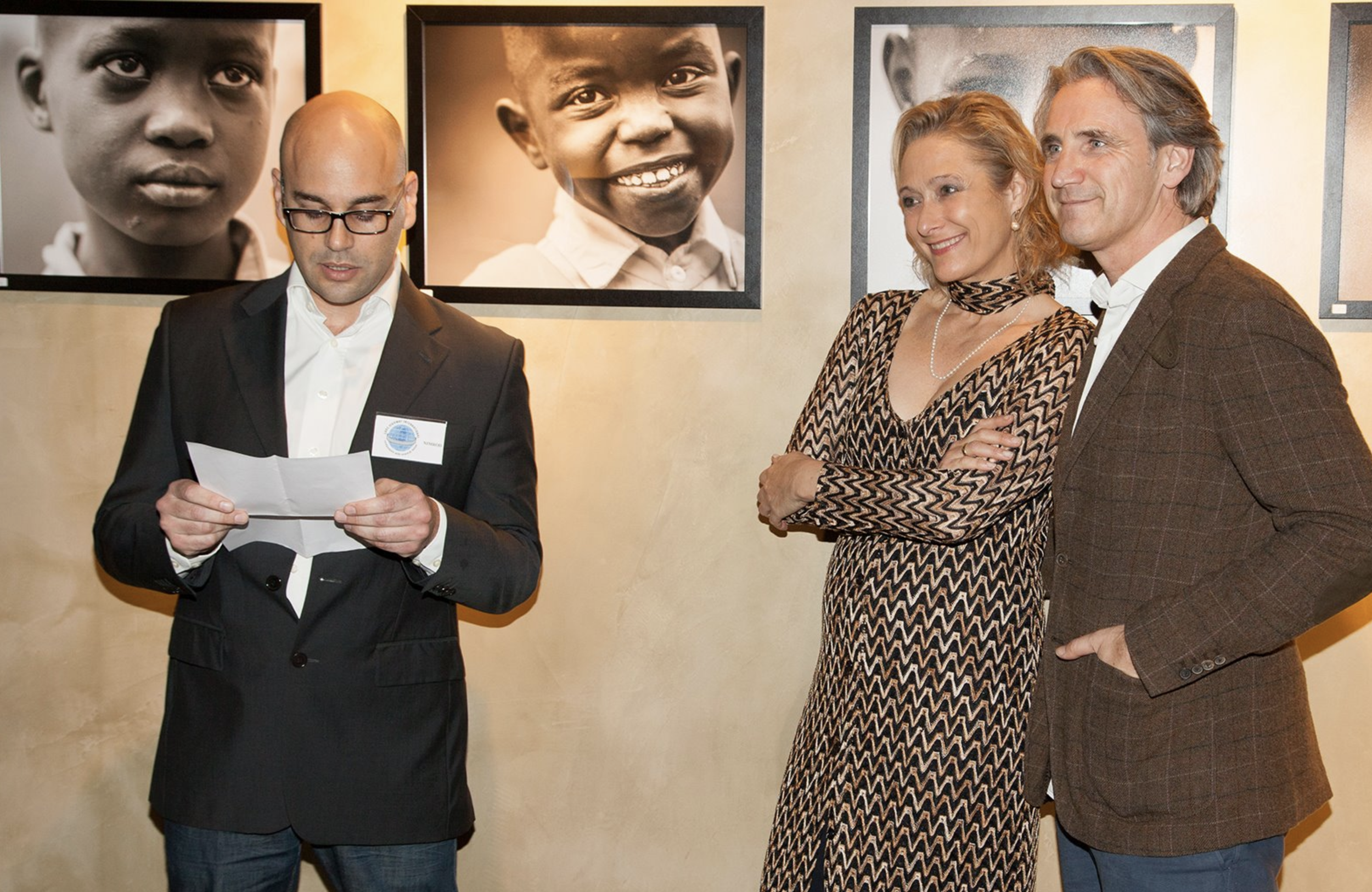 Man in black suit reading paper with two smiling women standing beside him at an art gallery with black and white portrait photographs on the wall behind them.