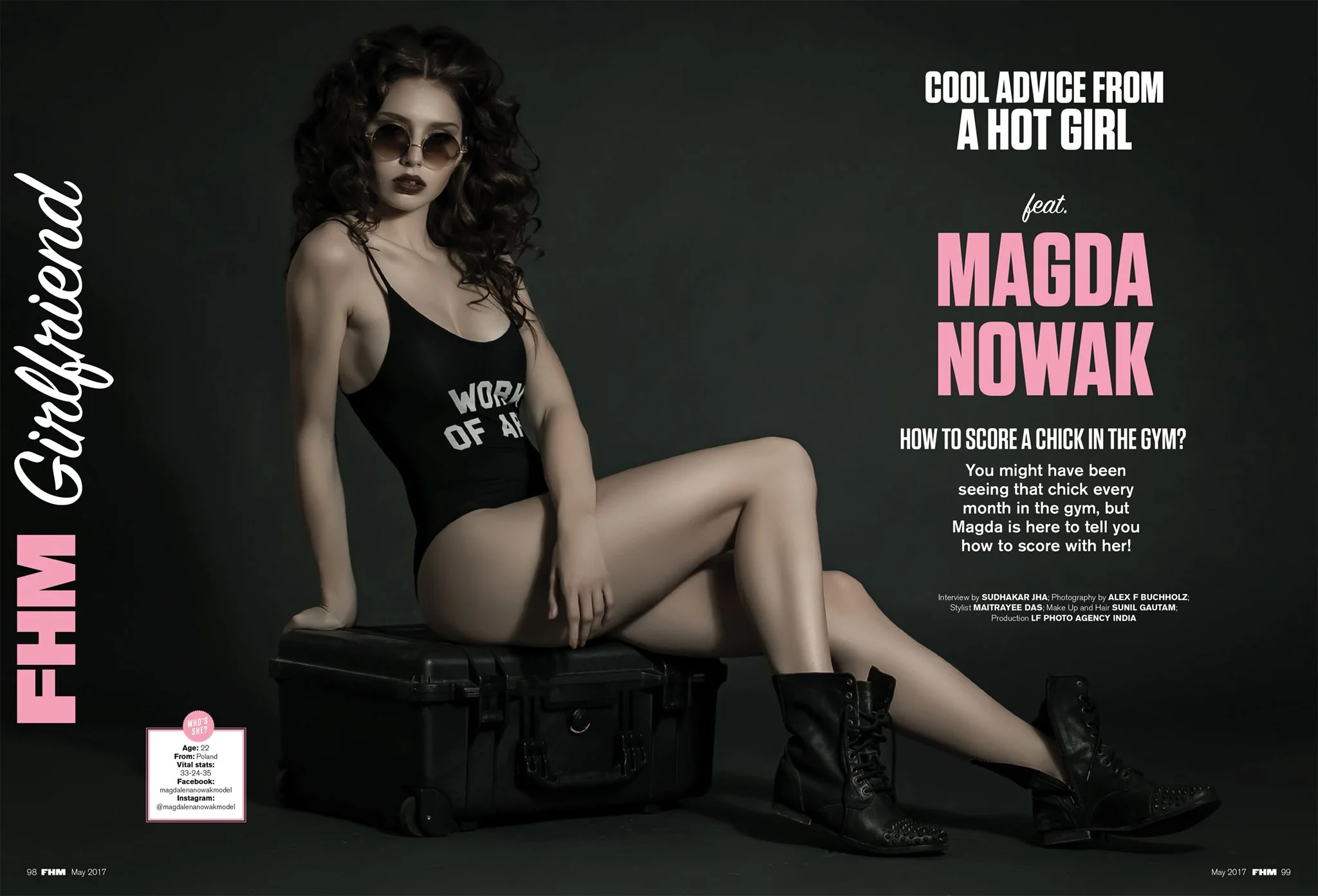 Woman sitting on a black case, wearing black sunglasses, a black bodysuit with 'World of Arms' written on it, black boots, with dark curly hair, posing against a dark background.