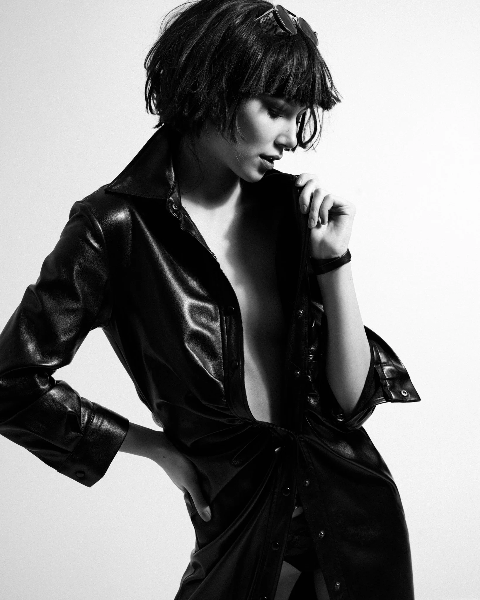A black and white photo of a woman with a short, choppy hairstyle wearing sunglasses on her head, leather jacket, and pants. She is posing with her head slightly bowed and eyes closed.