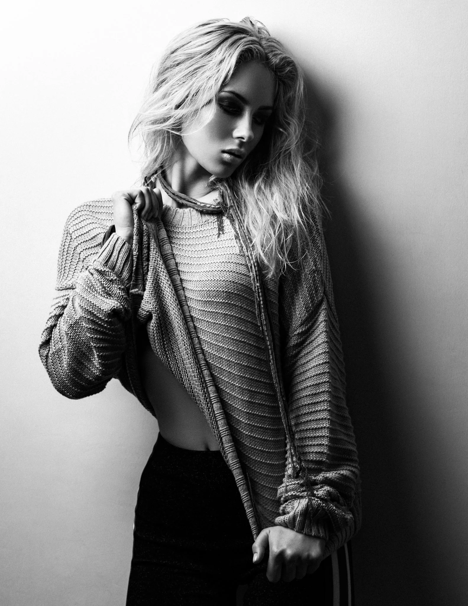 A young woman with wavy blonde hair, wearing a striped zip-up hoodie and black pants, is standing against a plain wall with her head tilted downward and eyes closed.