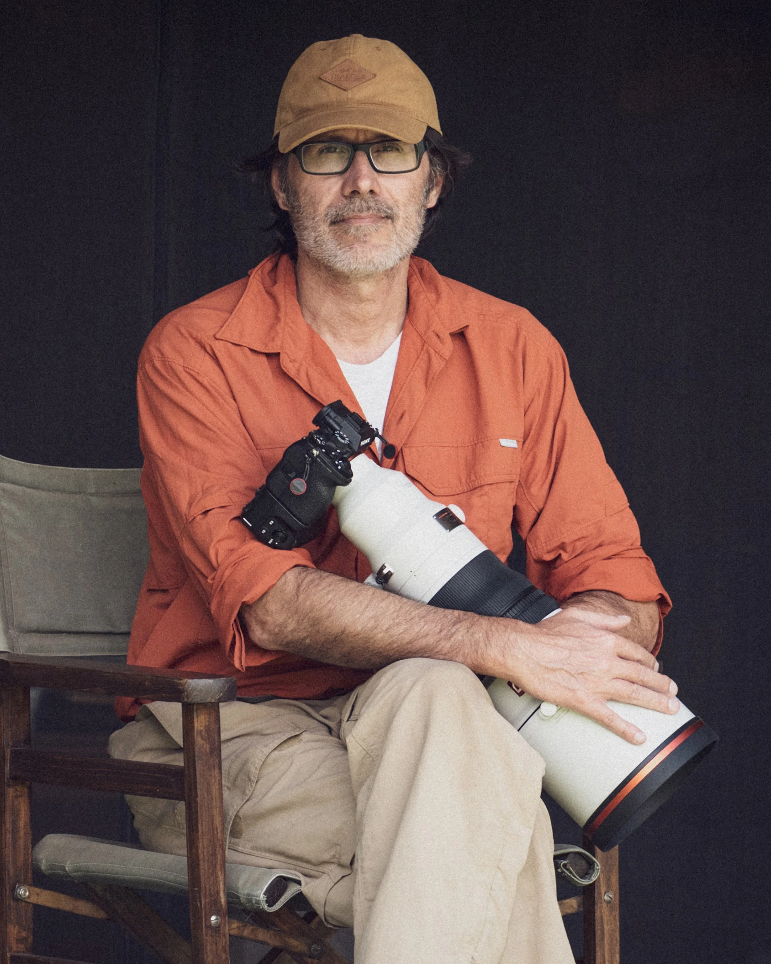 A man with glasses, a tan cap, orange shirt, and khaki pants sitting on a wooden chair, holding a large telephoto camera lens.