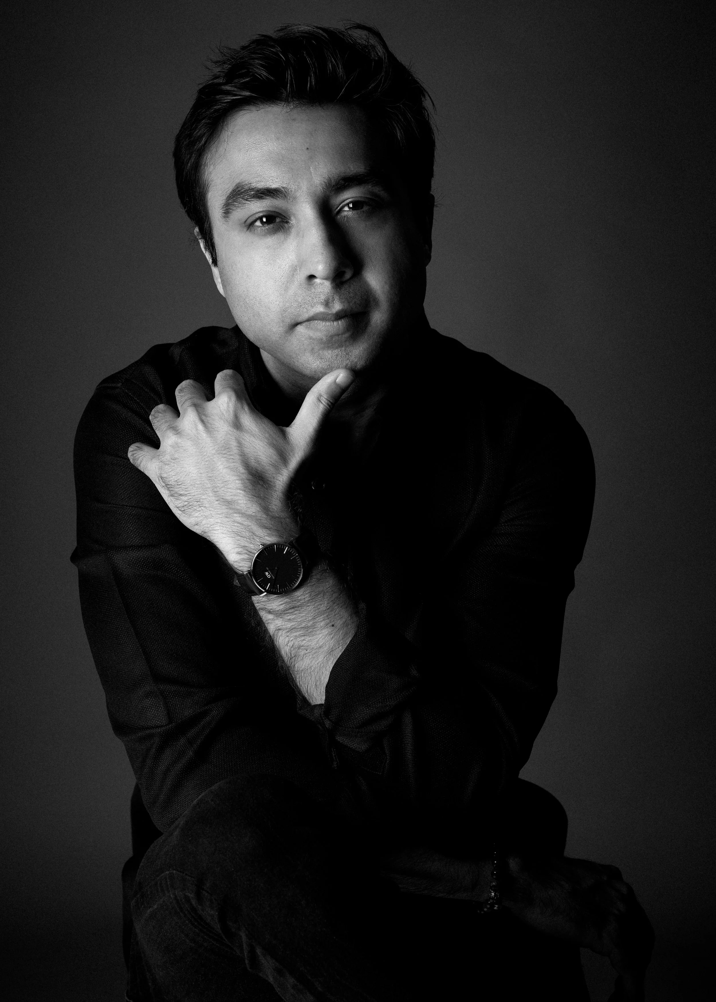 Black and white portrait of a man with dark hair, wearing a long sleeve shirt and wristwatch, resting his chin on his hand with a contemplative expression.