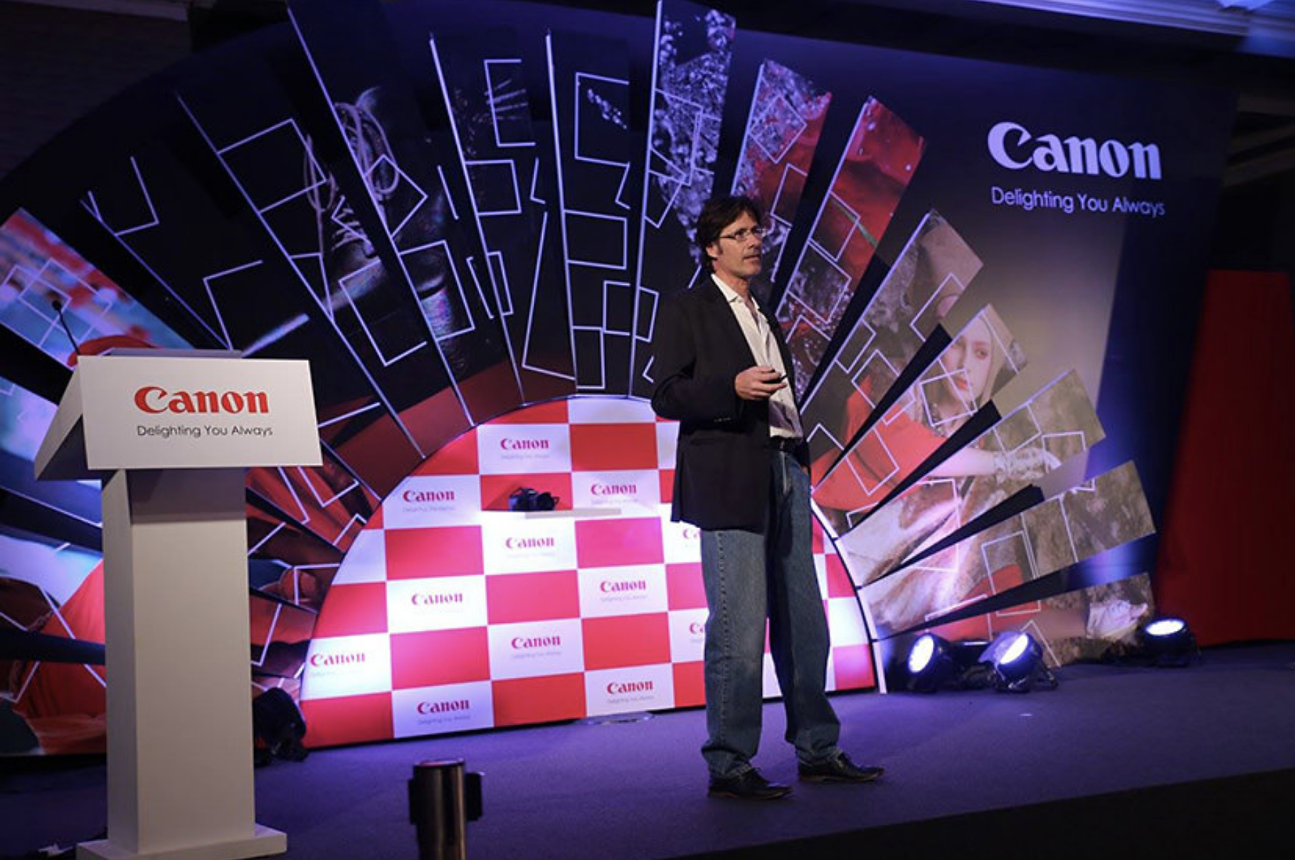 A man in a black blazer and jeans standing on stage with a Canon backdrop at a corporate event or presentation.