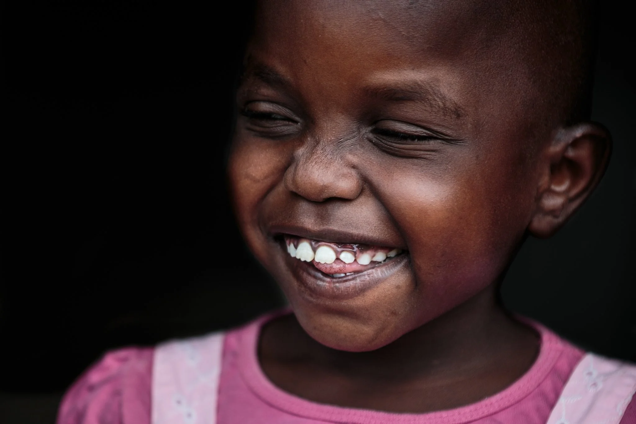 Close-up of a smiling young girl with dark skin, wearing a pink shirt, showing her teeth and a missing upper front tooth, against a dark background.